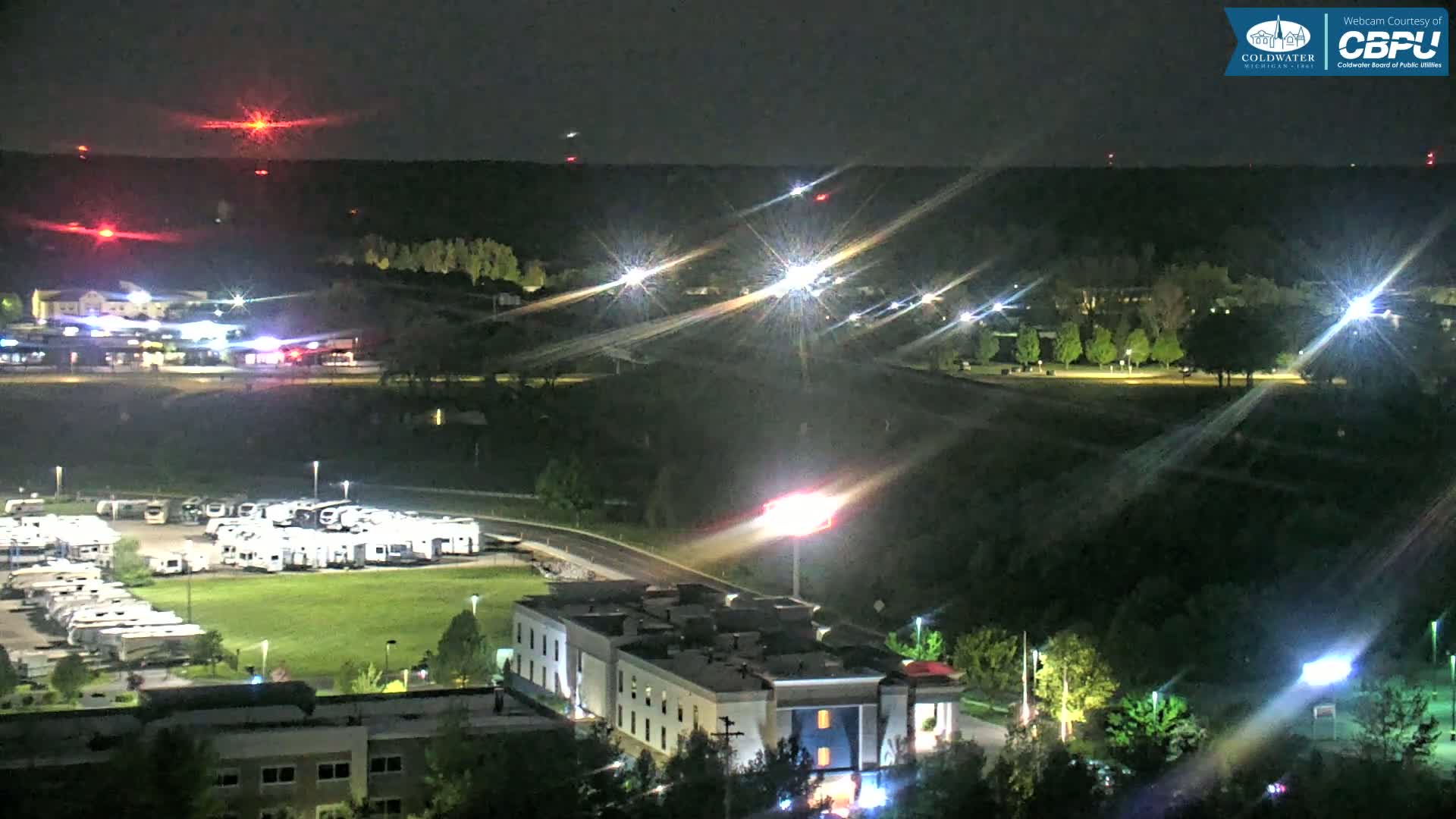 A nighttime, aerial view shows a parking lot full of RVs, a multi-story building, and a grassy area, all illuminated by artificial lights, with a dark, wooded area and distant red lights visible in the background.