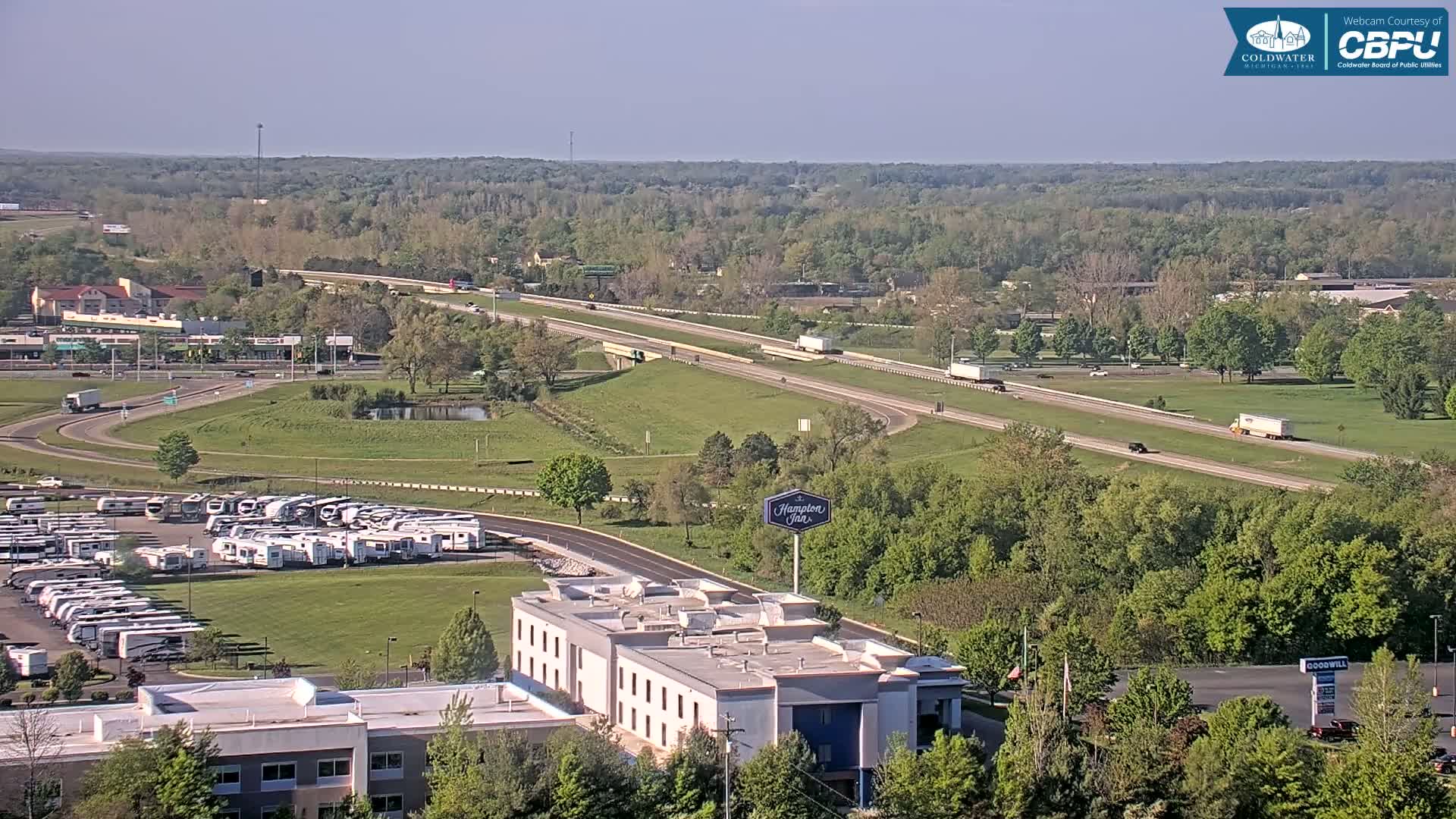 A mostly sunny aerial view shows a highway with several vehicles, a large RV park, a Hampton Inn, and a surrounding landscape of green trees and grass.