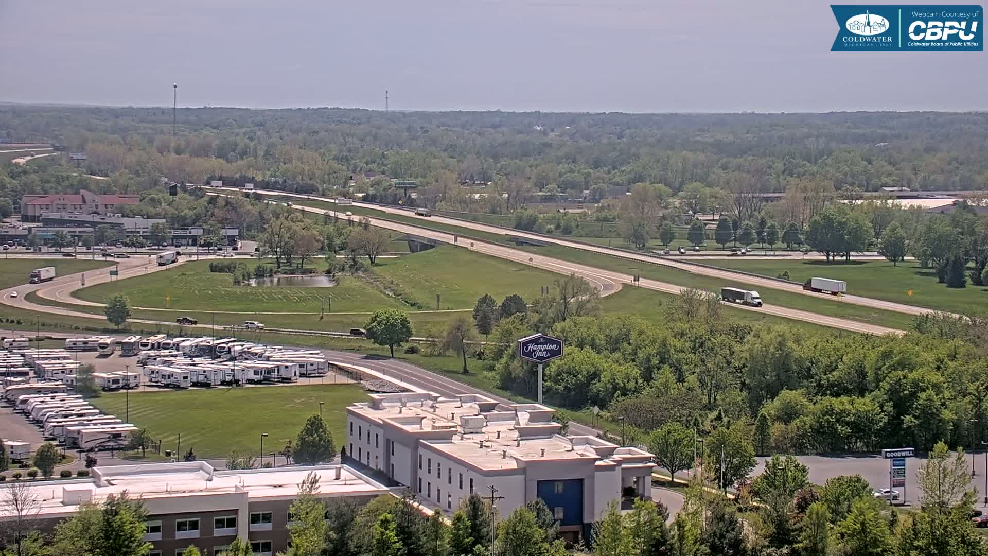 A high-angle, long shot on a sunny day shows a highway with several vehicles, a large RV park, a Hampton Inn, and surrounding green trees and buildings.