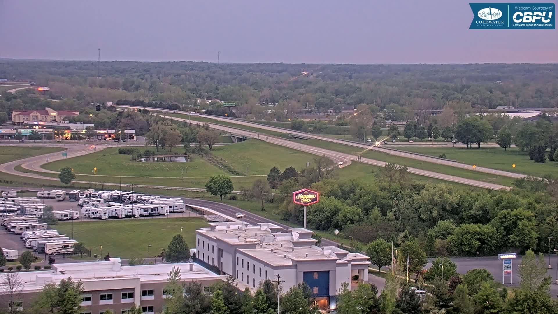 A high-angle, long shot shows a highway with sparse traffic, a large RV park, a hotel, and a mostly-wooded area under a twilight sky.