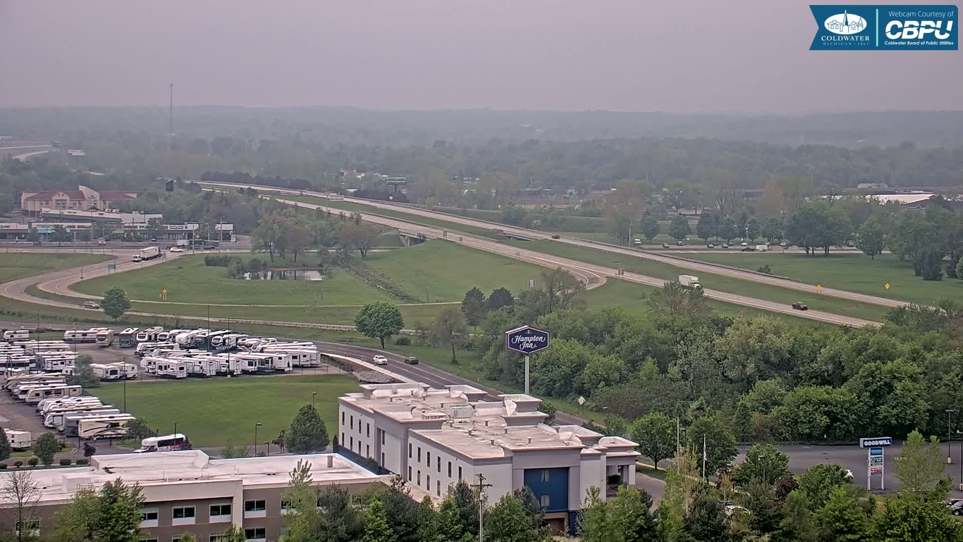 A hazy view of a highway, a large RV park, a Hampton Inn, and surrounding green space with many trees.