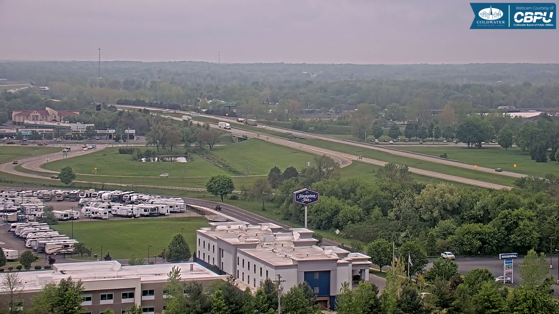 A hazy, overcast day reveals a highway flanked by green trees and a parking lot full of RVs near a Hampton Inn.