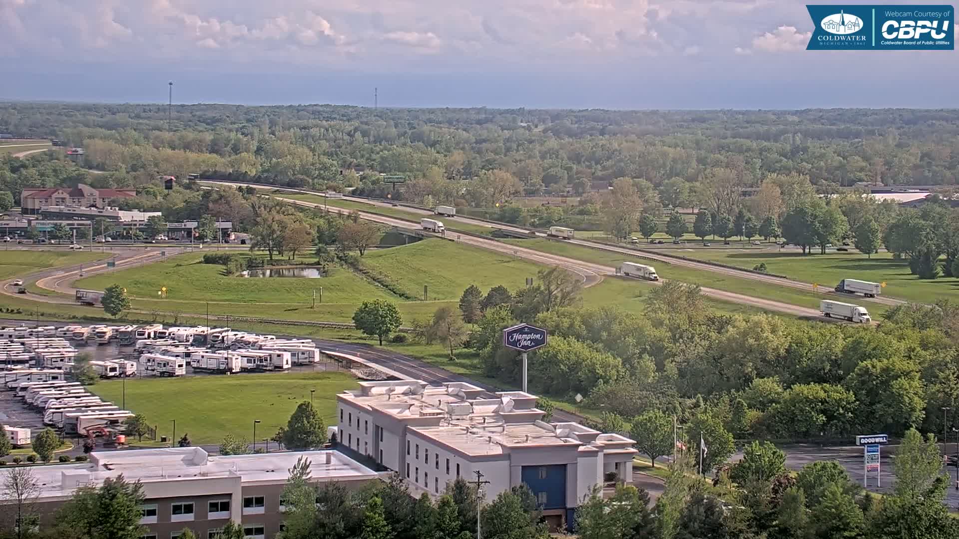A partly cloudy day shows a highway with several semi-trucks driving on it, alongside a large RV park and a Hampton Inn, situated within a green, tree-lined area.