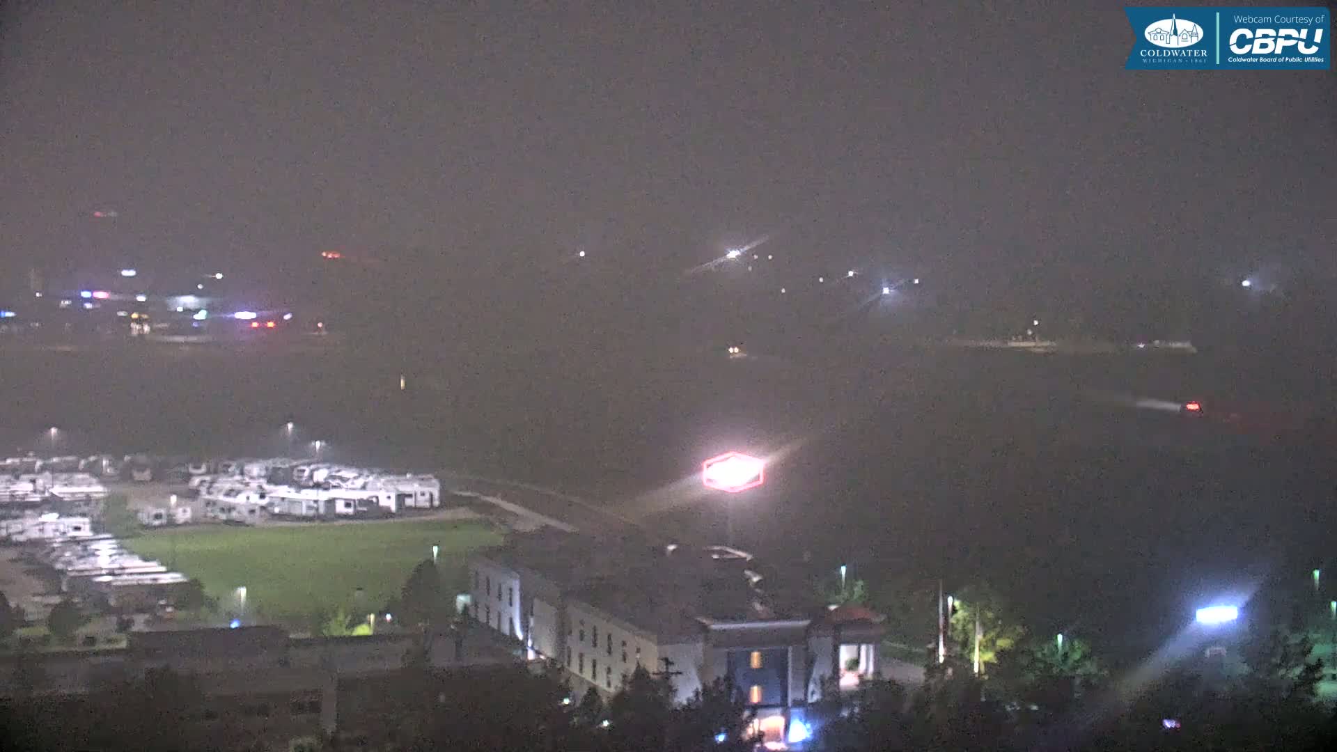 A hazy night scene shows a cluster of RVs, a hotel, and distant lights across a body of water.