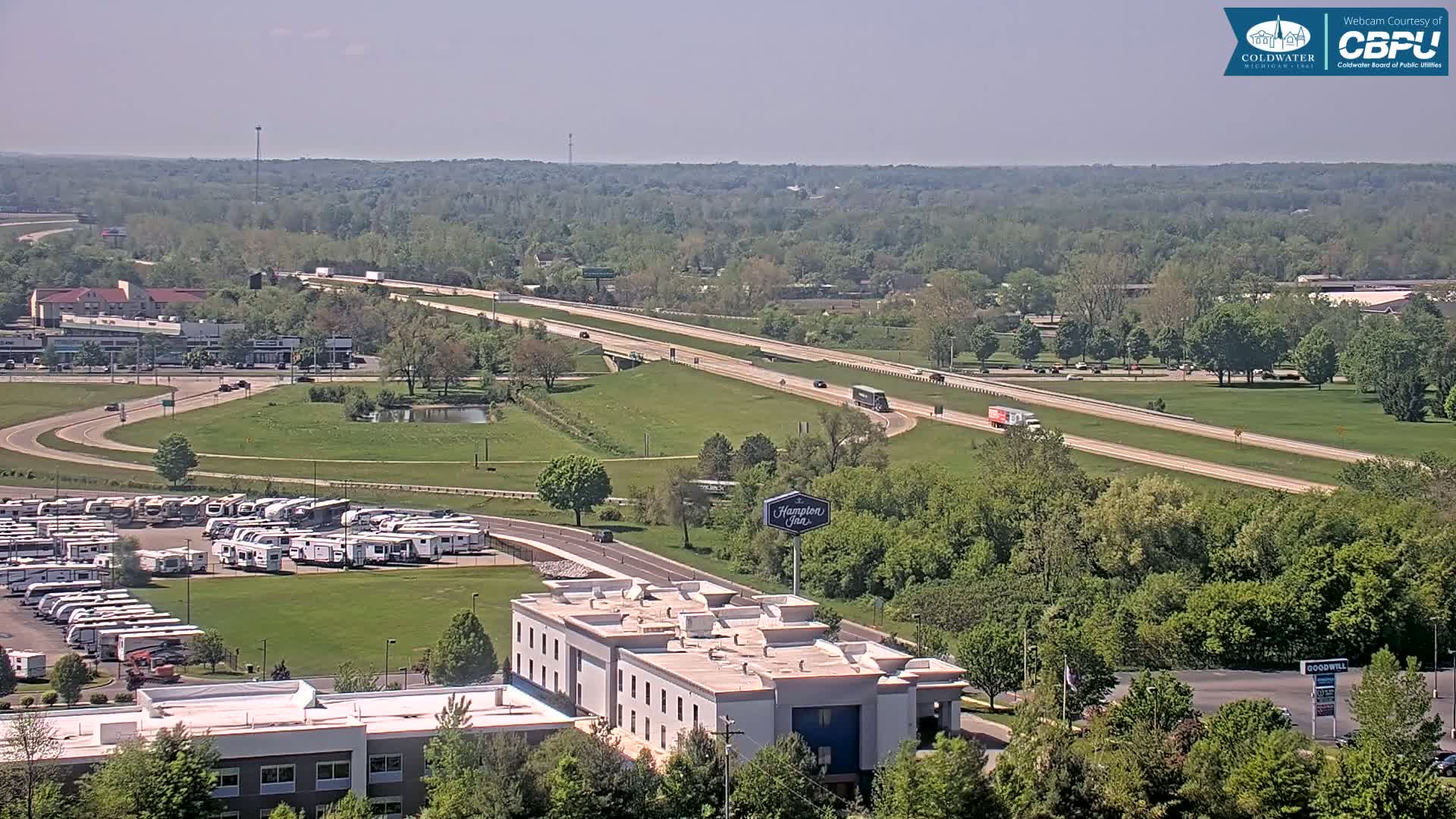A high-angle, long shot on a clear day shows a multi-lane highway passing through a green landscape, with a large parking lot full of RVs next to a hotel and other businesses.