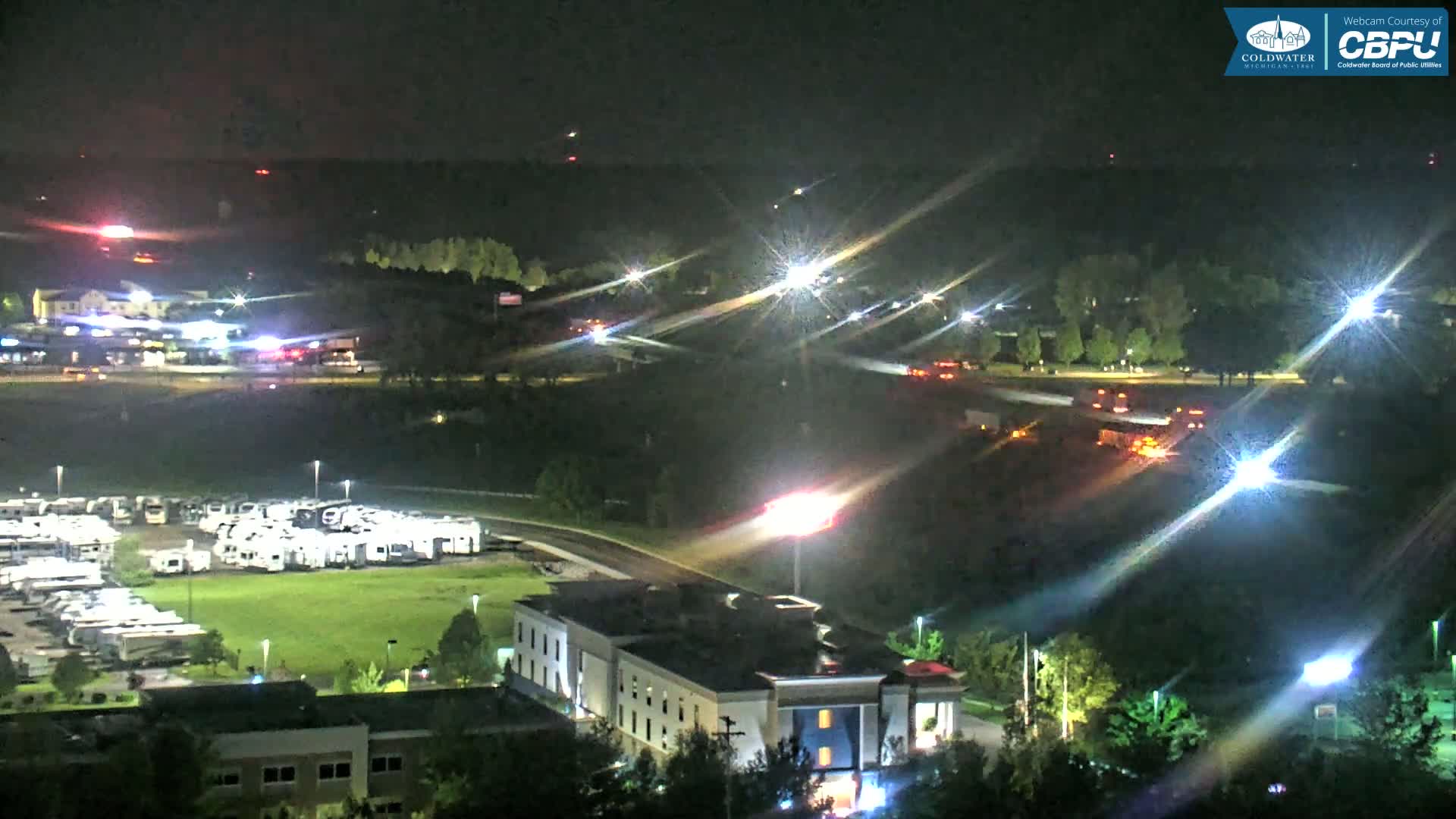A nighttime aerial view shows a large RV park, a hotel, and brightly lit roads with some vehicle traffic under a clear, dark sky.
