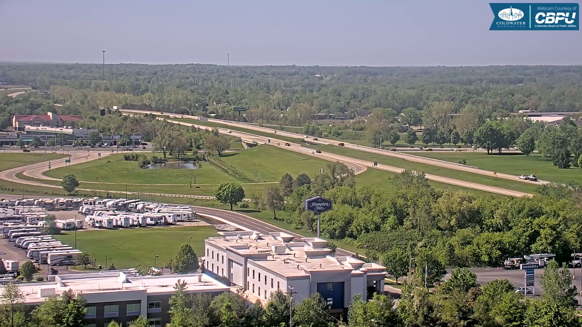 A highway with sparse traffic runs through a verdant landscape under a clear, sunny sky, alongside a parking lot filled with RVs and a Hampton Inn hotel.
