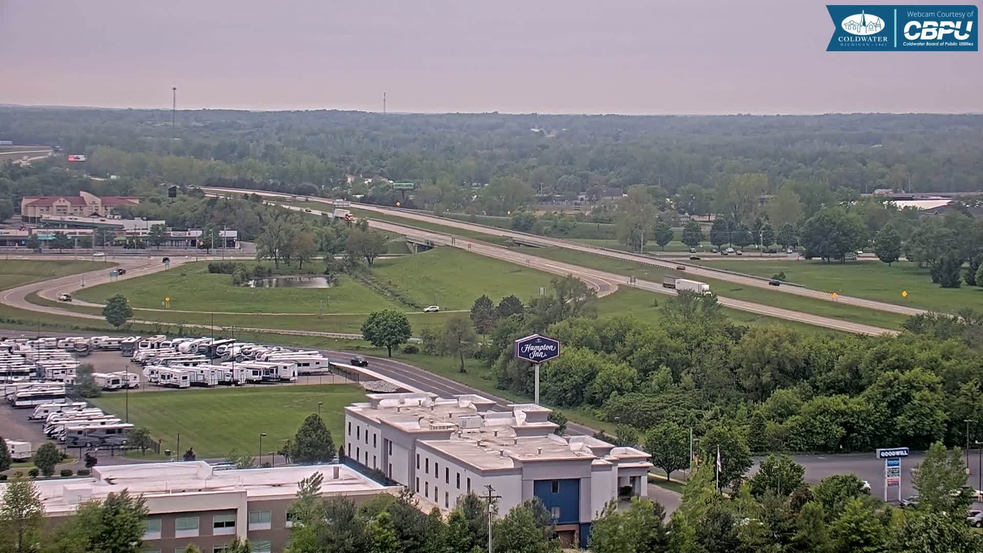 An aerial view shows a highway with light traffic passing through a lush green landscape, next to a parking lot filled with RVs and a Hampton Inn hotel.