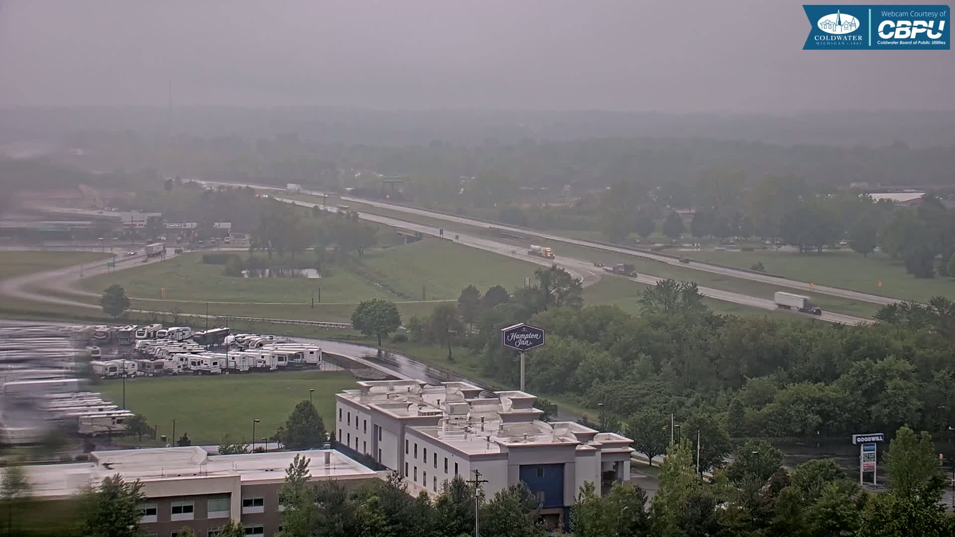 A hazy, rainy view shows a highway with several vehicles, a large parking lot filled with RVs, and a Hampton Inn, surrounded by green trees and grassy areas.