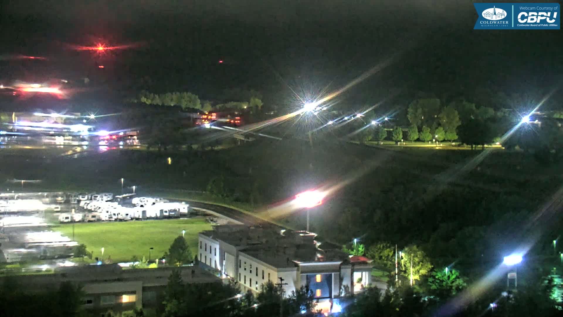 A nighttime aerial view shows a motel, a large parking lot filled with RVs, and a road with some vehicle lights visible, all under a dark, clear sky.