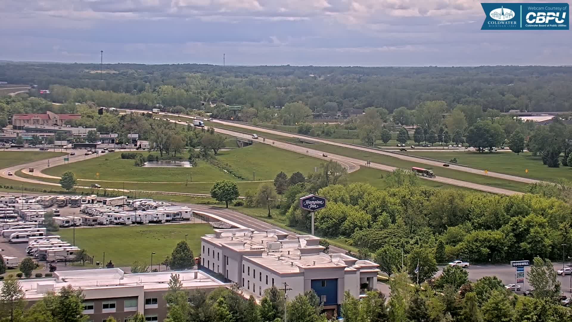 A highway with moderate traffic cuts through a green landscape dotted with buildings, including a Hampton Inn and a large RV park, under a partly cloudy sky.