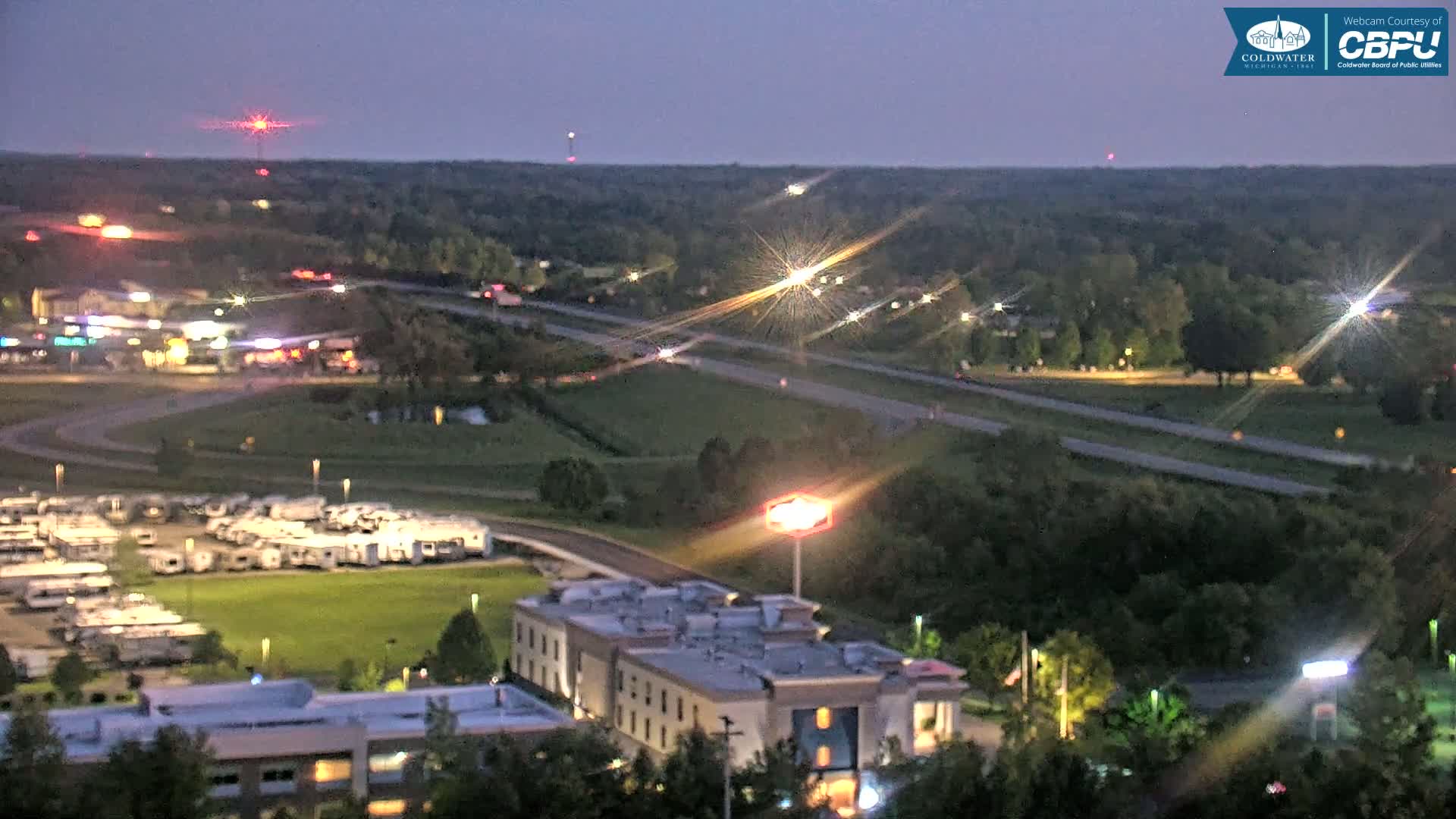 A twilight view shows a highway, a large RV park, a hotel, and a sprawling wooded area, all illuminated by artificial lights under a dusky sky.