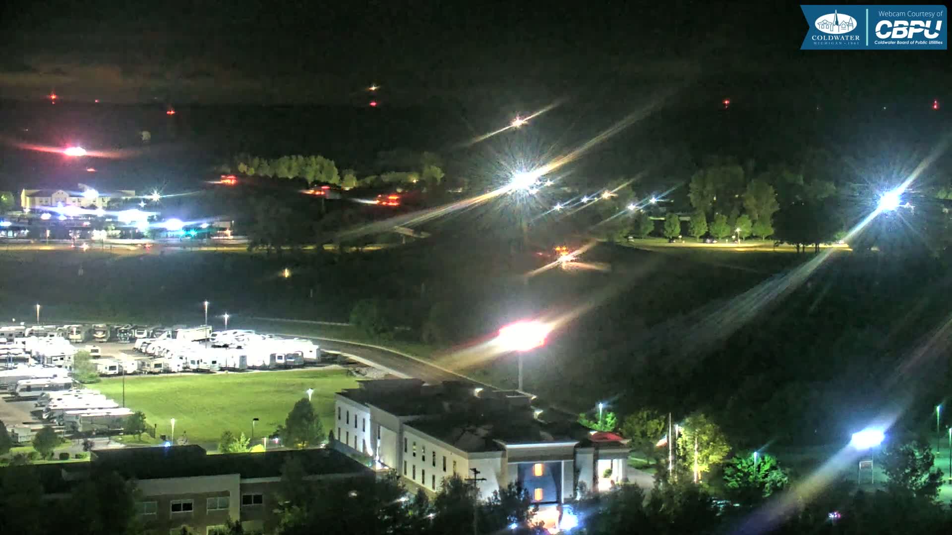 A nighttime, high-angle view shows a large parking lot filled with RVs, a hotel, and surrounding trees and roads illuminated by artificial lights.