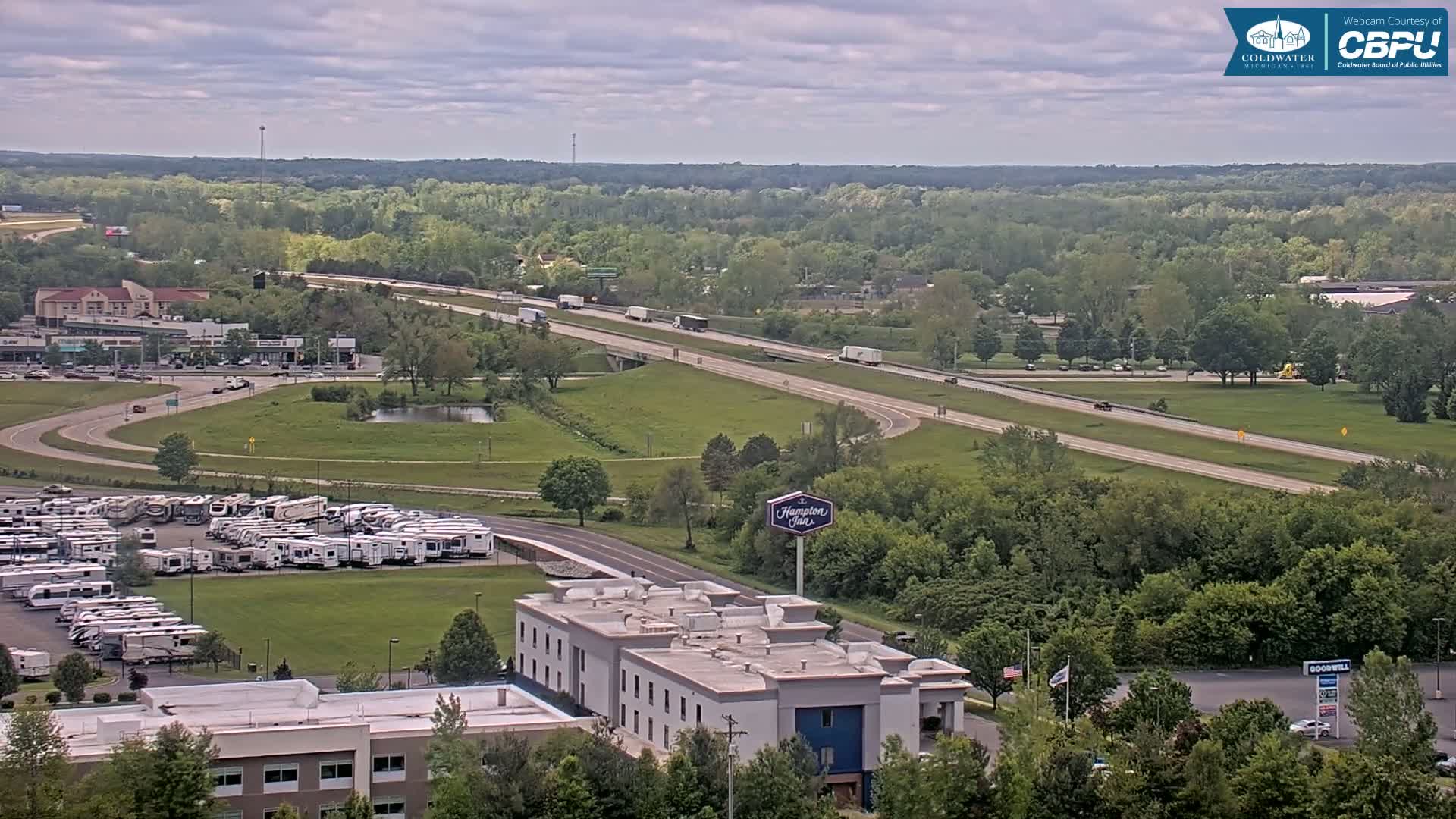 An aerial view on an overcast day shows a highway with several vehicles, a large RV park, a Hampton Inn, and a sprawling green landscape dotted with trees.