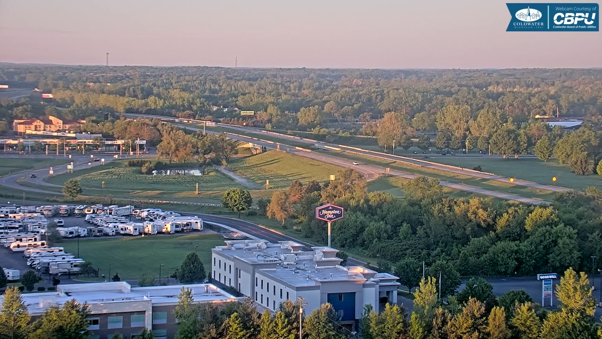 A high-angle, long shot of a highway, a Hampton Inn, an RV park, and a large expanse of green trees under a clear, pale sunrise sky.