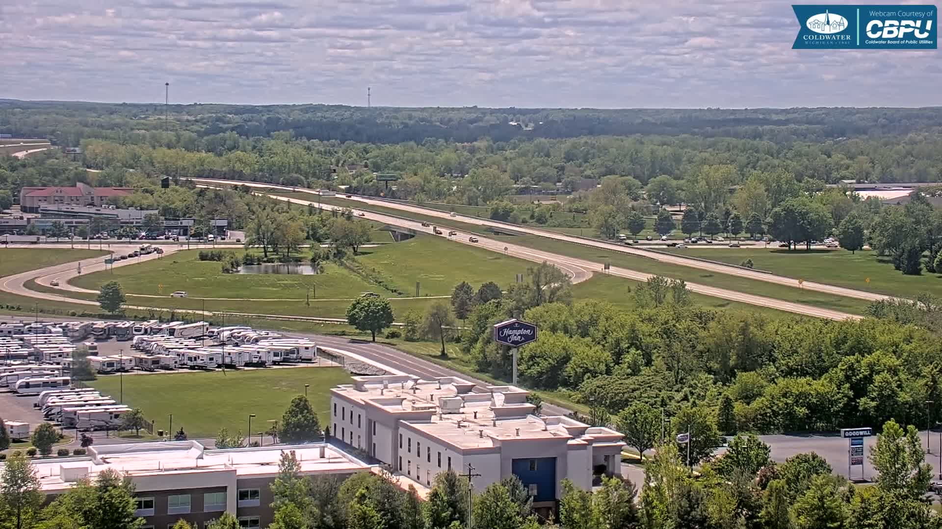 A partly cloudy day reveals a highway cutting through a green landscape with a large RV park, a hotel, and other buildings nearby.