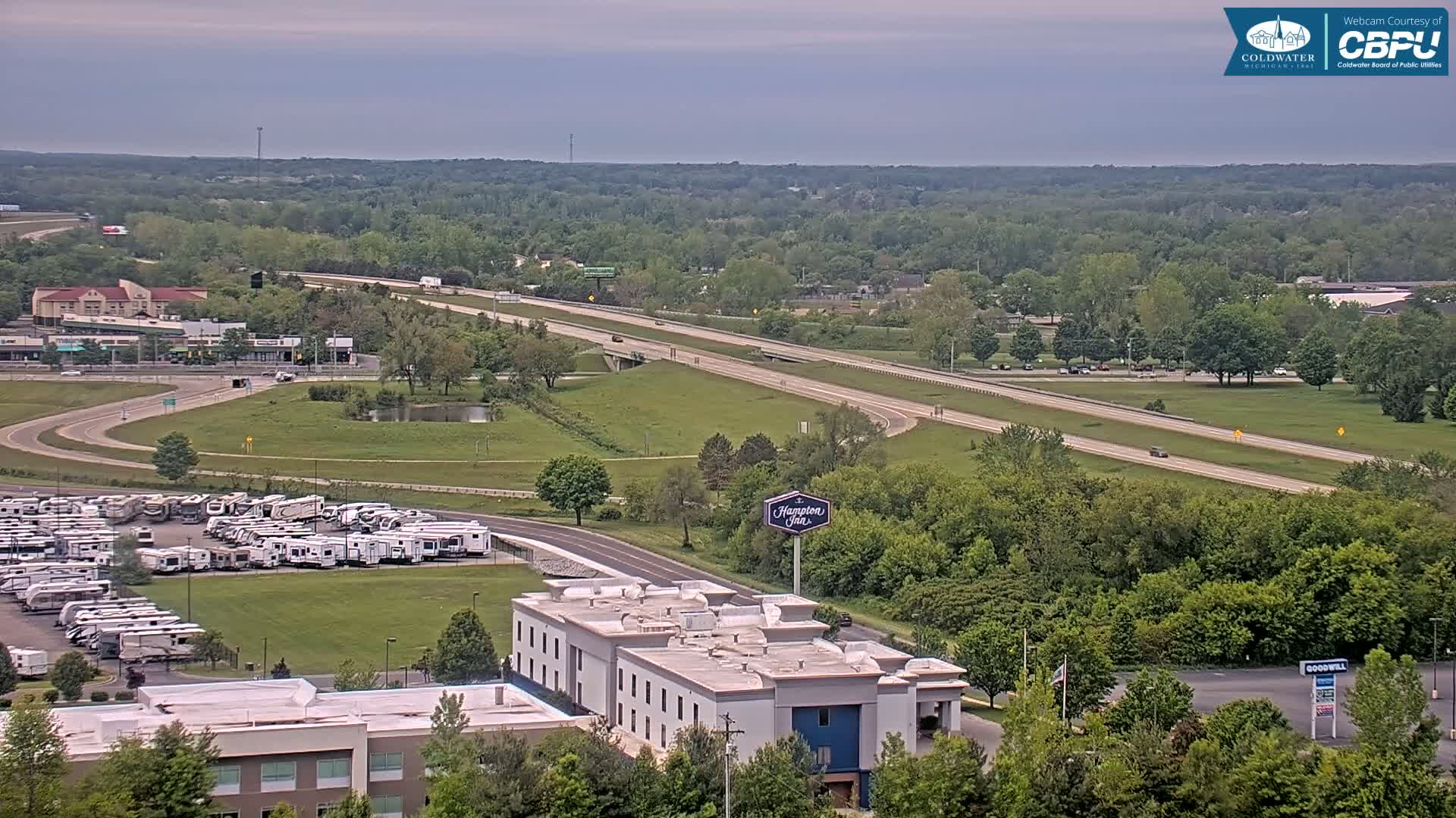 A wide shot shows a highway and surrounding area under overcast skies, featuring a large RV park, a Hampton Inn, and green, leafy trees.
