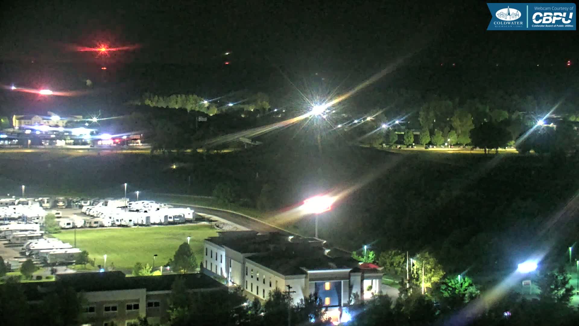 At night, a large RV park and a hotel are illuminated, with lights from vehicles and distant structures visible under a clear, dark sky.