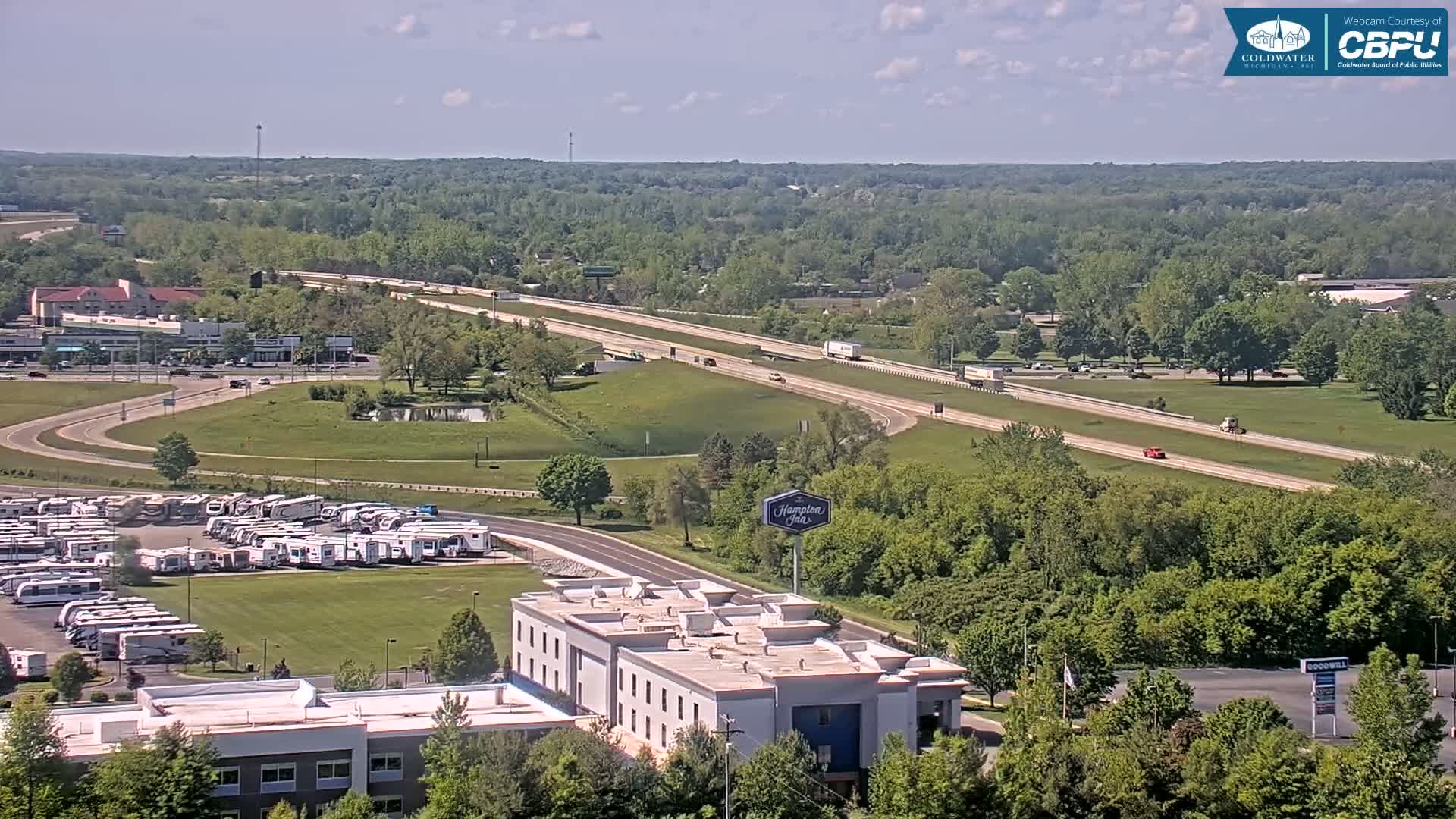 A highway with sparse traffic runs through a green landscape under a mostly sunny sky, alongside a large RV park and a hotel.