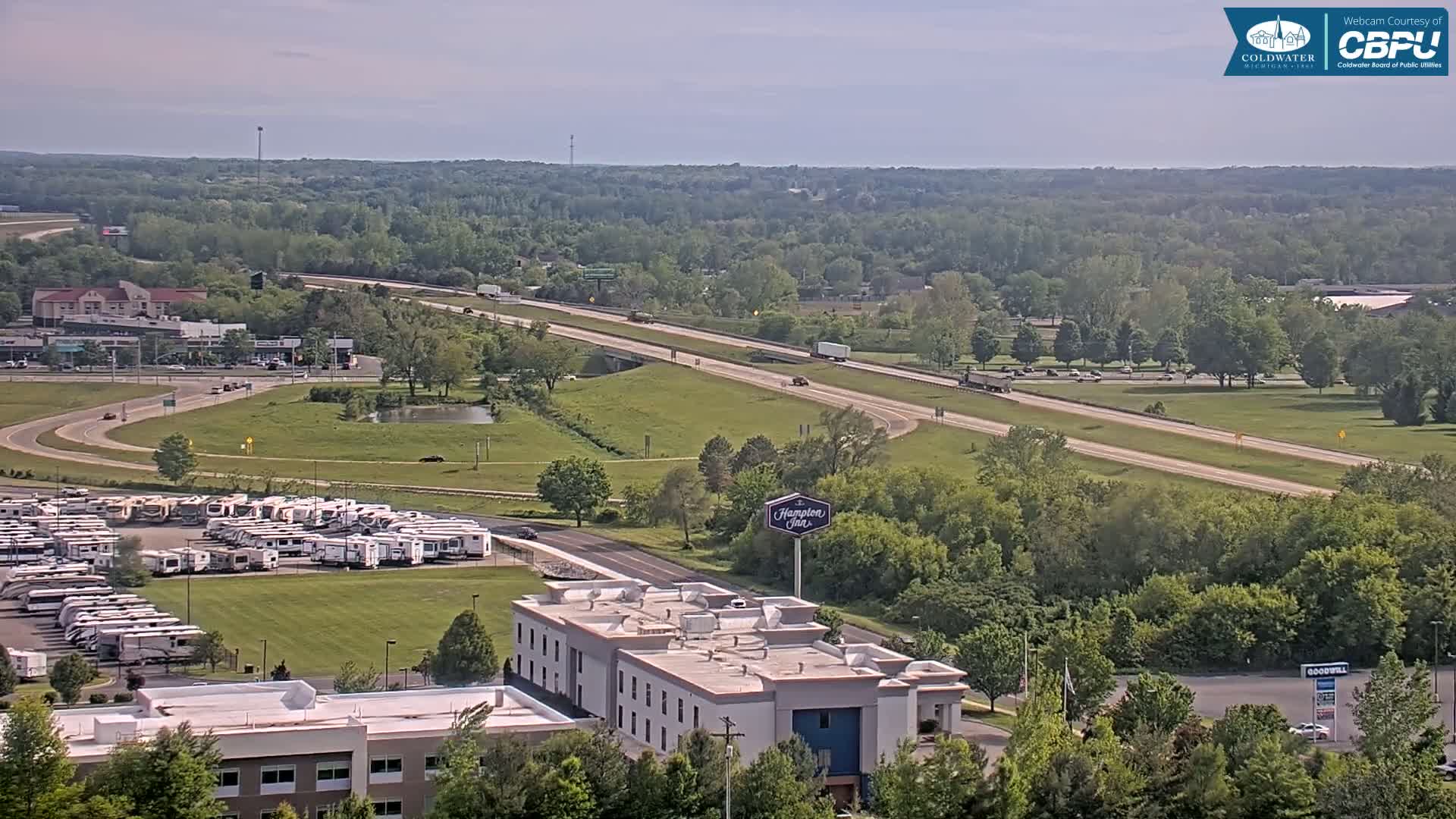 A high-angle, long shot on a partly sunny day shows a highway with several vehicles, a large RV park, a Hampton Inn, and surrounding green trees and buildings.