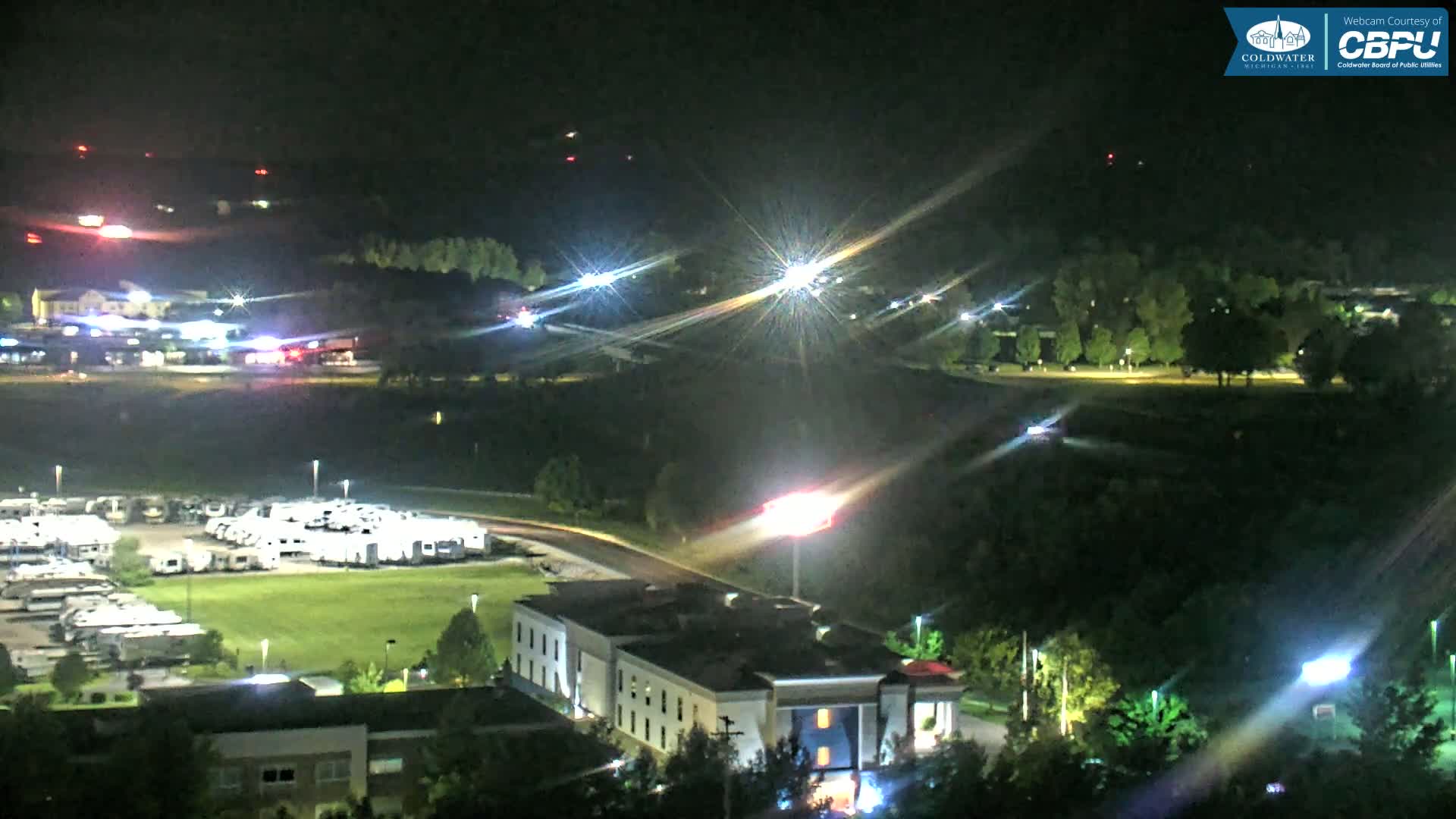 A nighttime aerial view shows a motel, a large RV park, and surrounding areas illuminated by lights, with some distant lights possibly indicating emergency vehicles.