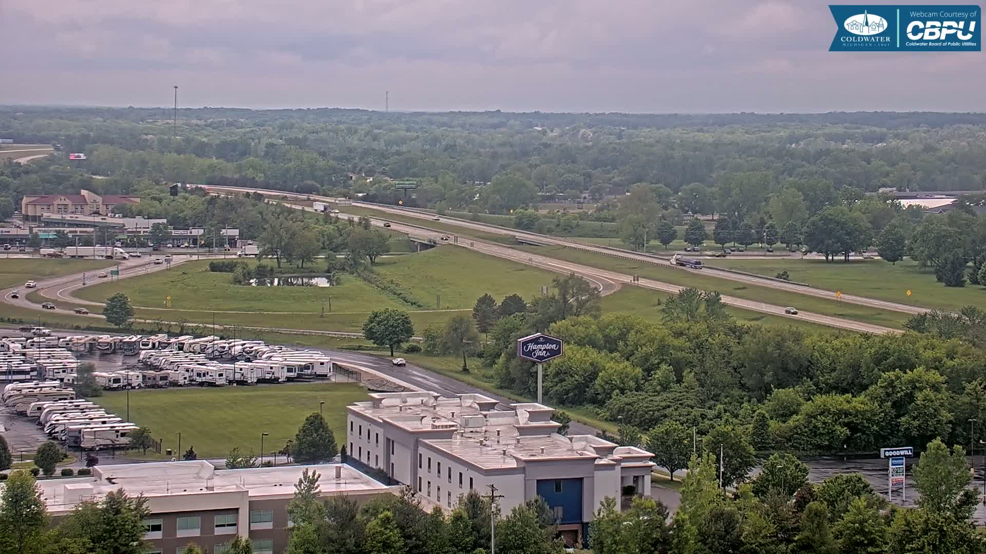 A high-angle view shows a highway, a large parking lot filled with recreational vehicles, a Hampton Inn, and a mostly overcast sky over a green, wooded landscape.