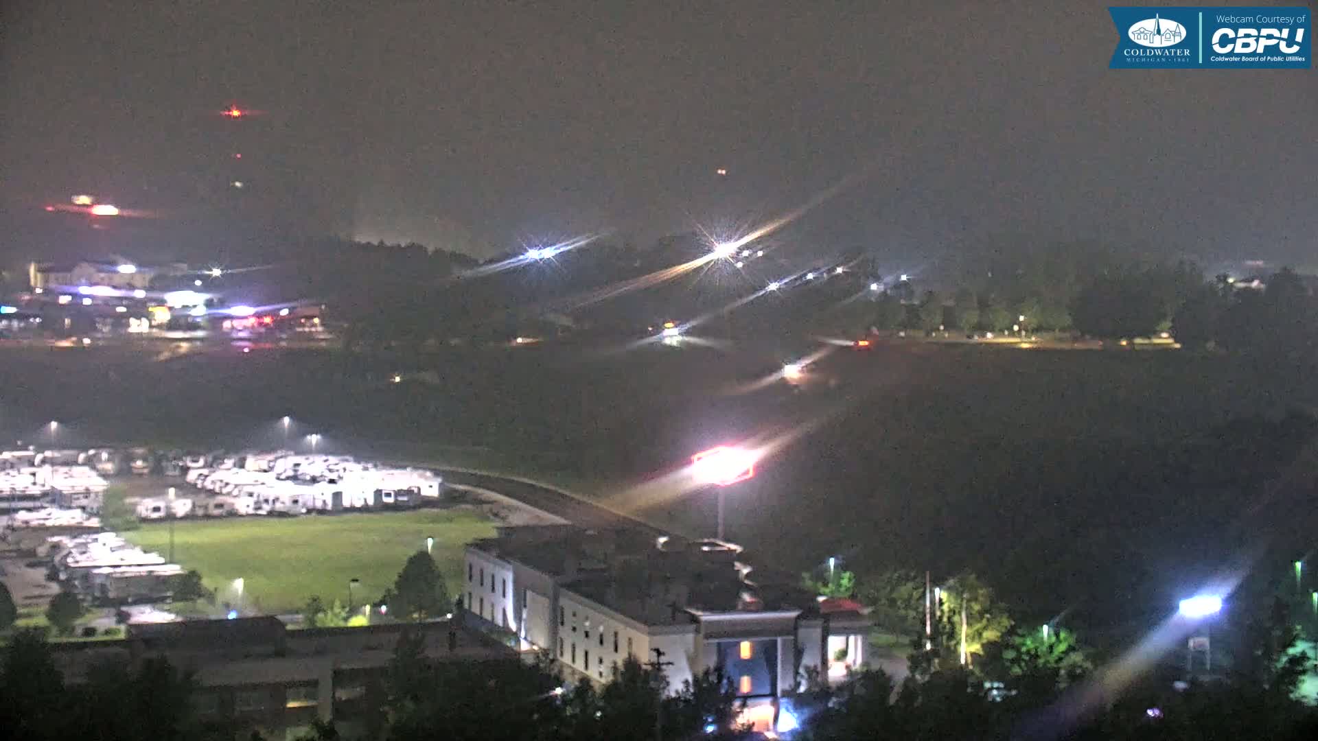 A nighttime, hazy view shows a large parking lot filled with RVs, a multi-story building, and a road with scattered cars and lights in the distance.