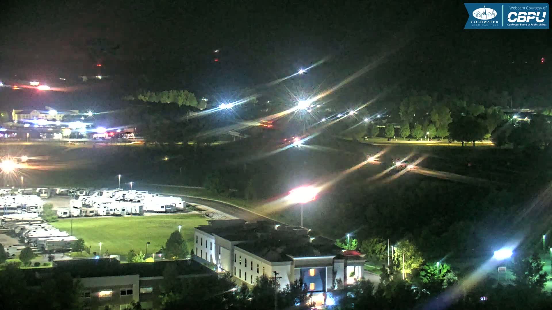 A nighttime aerial view of a highway intersection, a large RV park, and a hotel, with some vehicle traffic visible under a clear, dark sky.