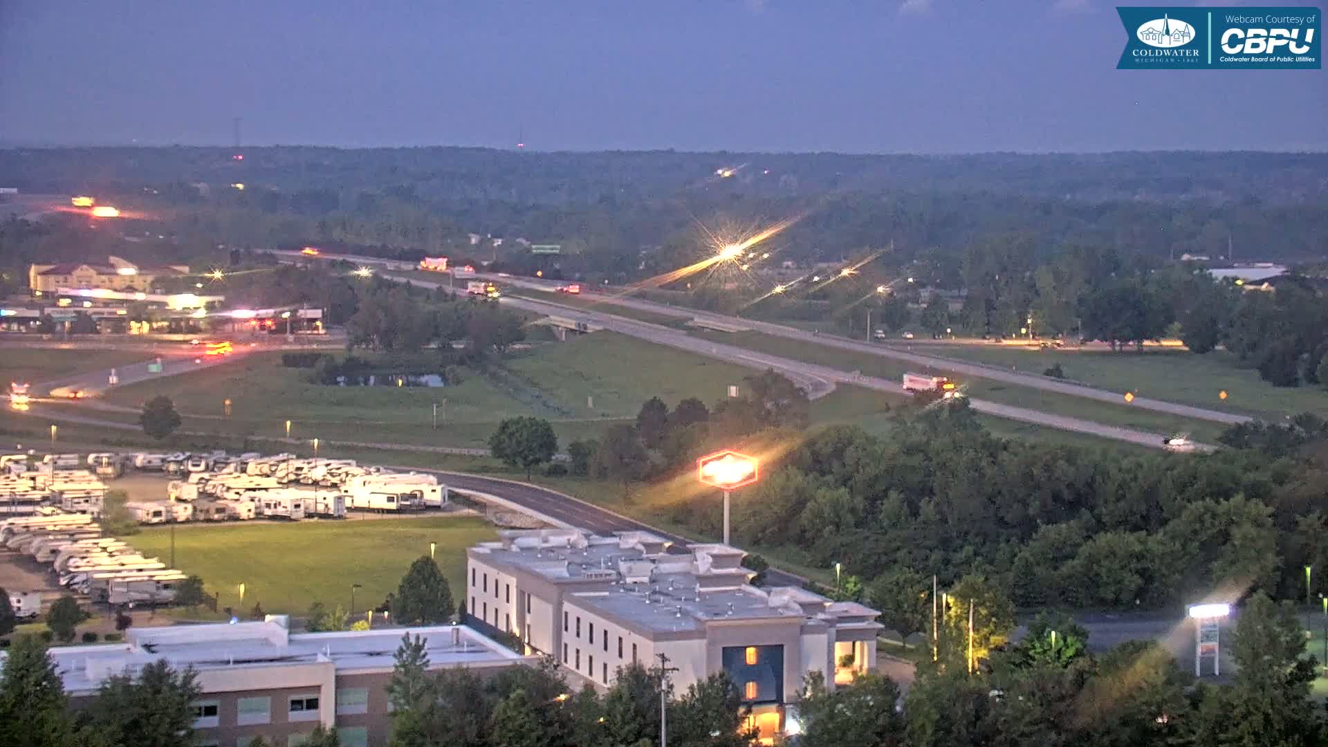 An elevated view at dusk shows a highway with several vehicles, a large RV park, a hotel, and surrounding green spaces under a clear, twilight sky.