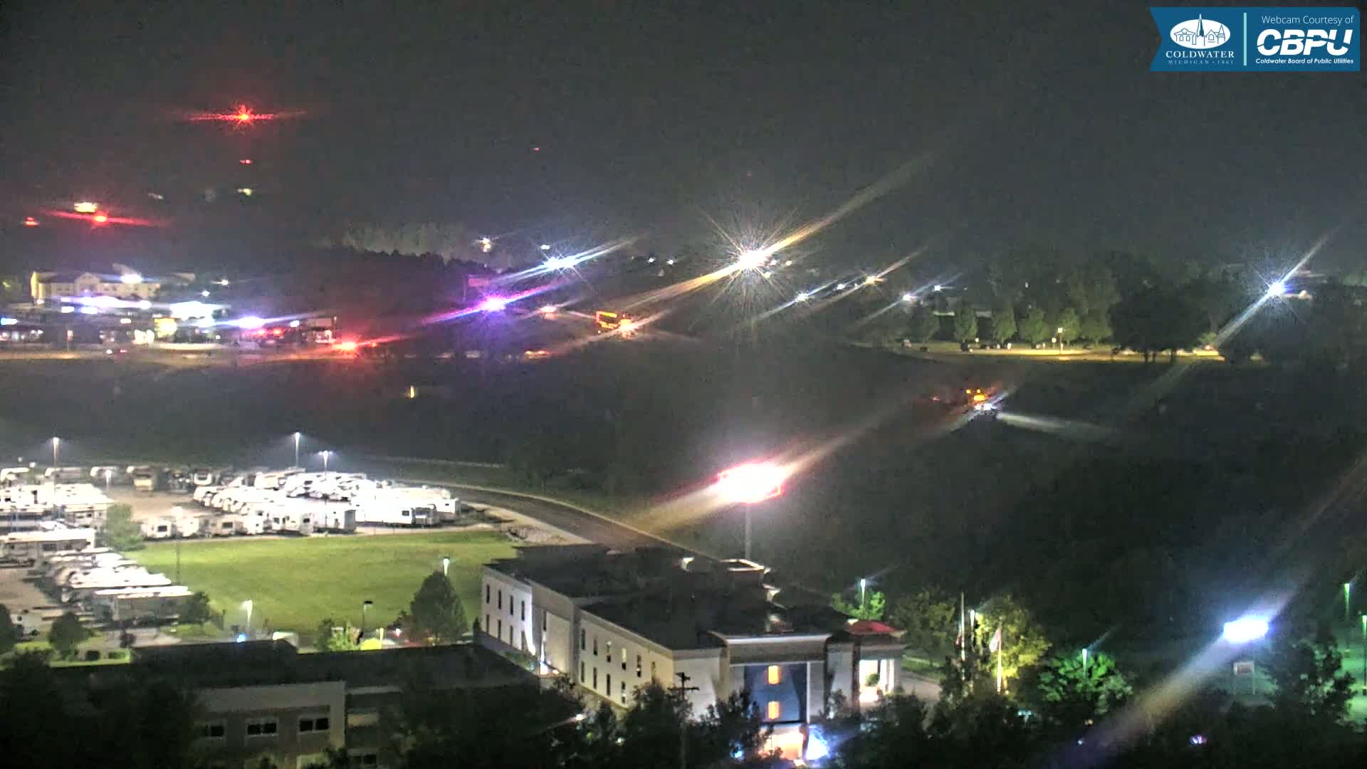 At night, emergency vehicles and flashing lights are visible in the distance, near a large parking lot full of RVs and a hotel.