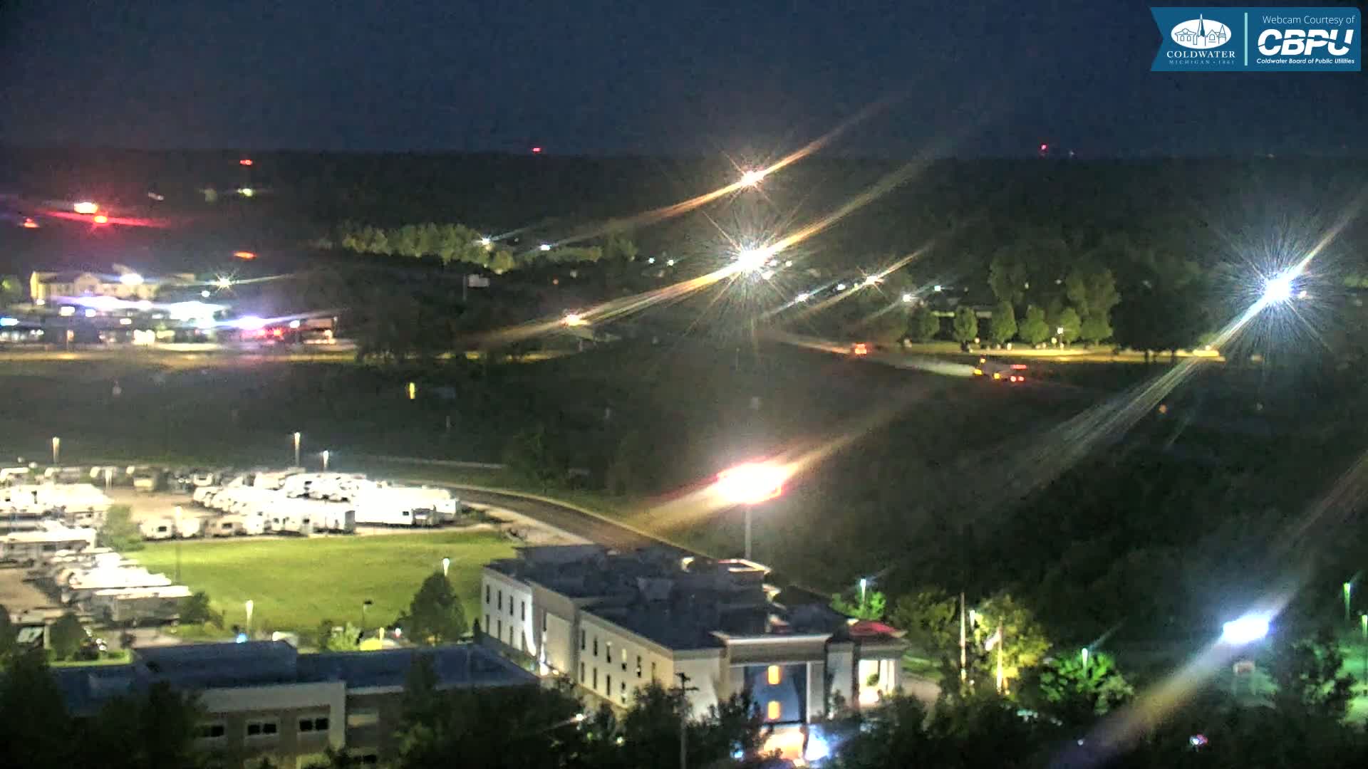 A nighttime aerial view shows a parking lot full of RVs, a multi-story building, and a roadway with some vehicles, all surrounded by dark, tree-covered hills.