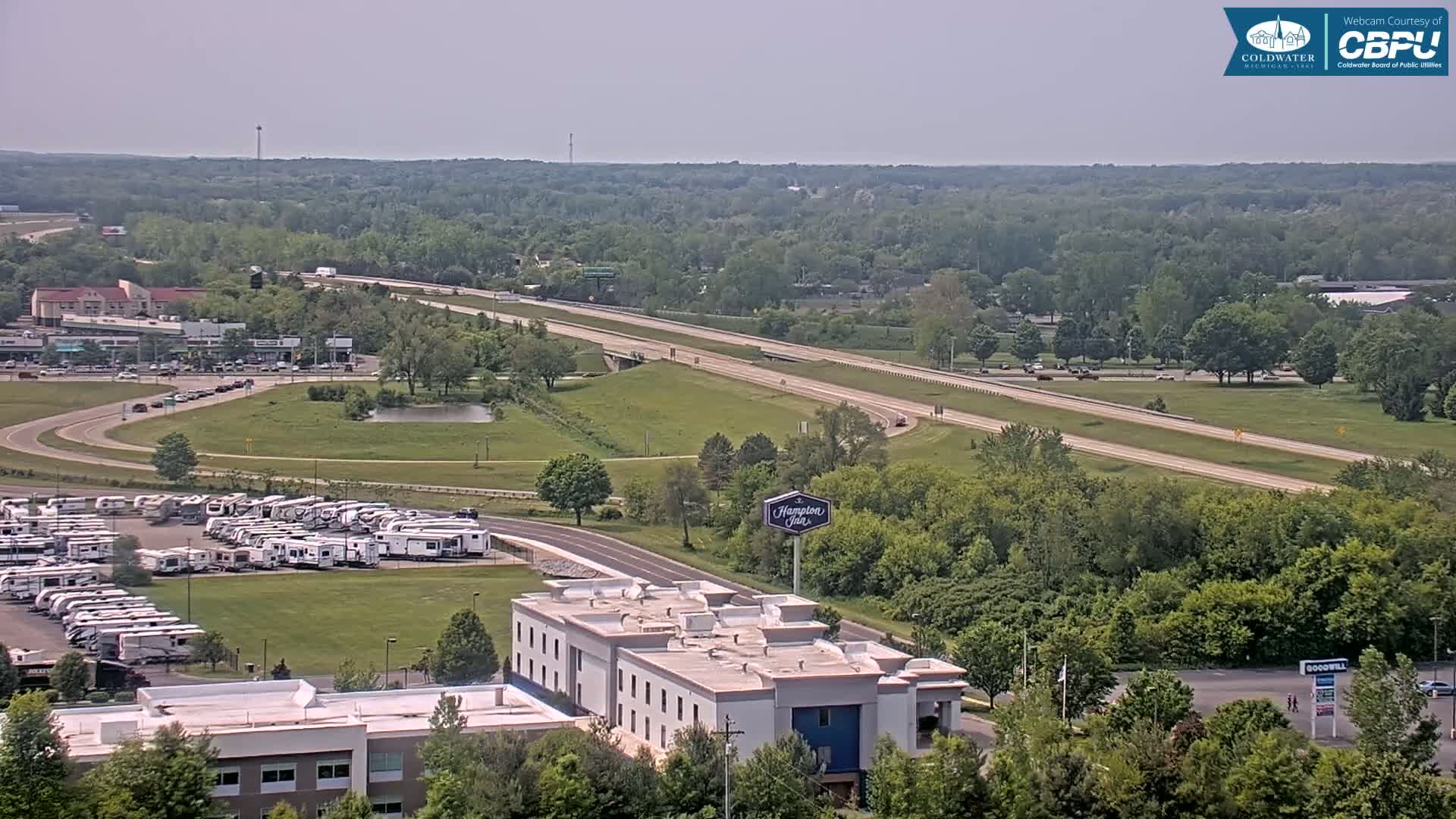 A high-angle, long shot on a sunny day shows a highway and surrounding area with a Hampton Inn, a large RV park, and green, leafy trees.