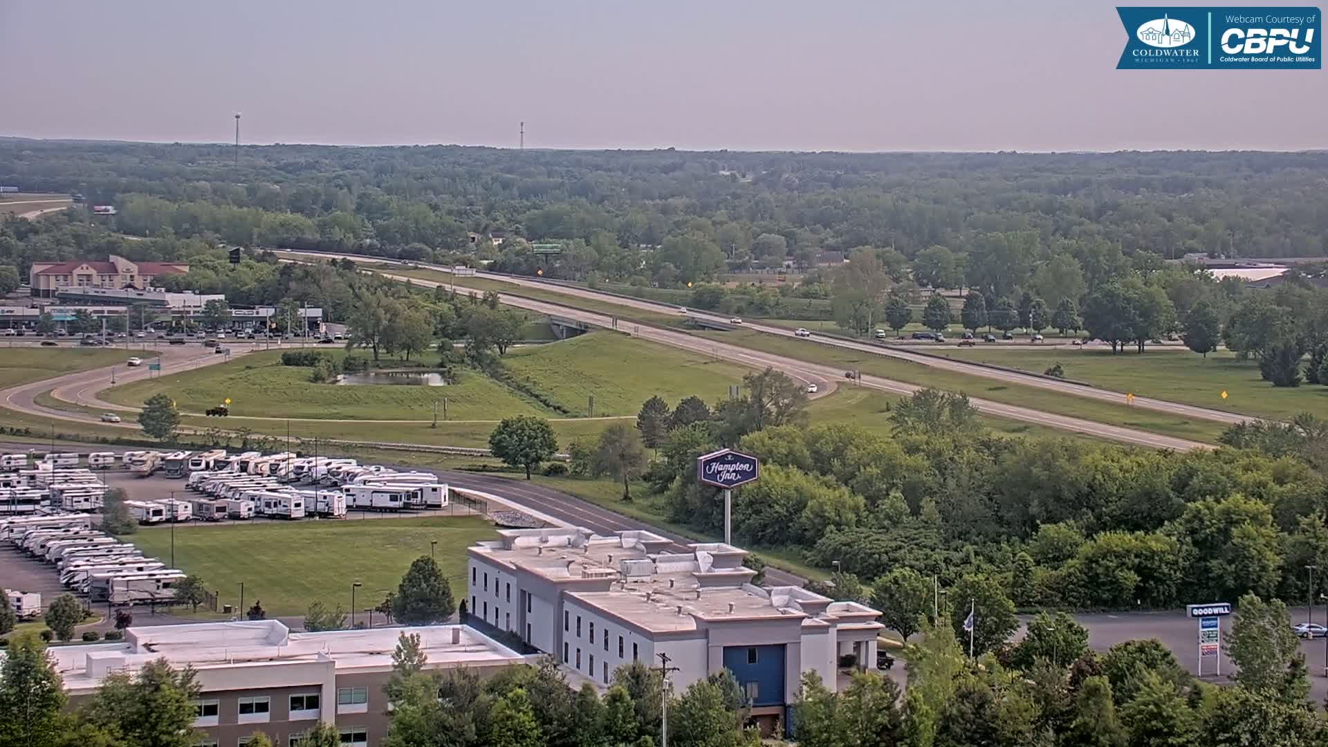A mostly sunny day shows a highway and a large RV park near a Hampton Inn, surrounded by green trees and a few other buildings.