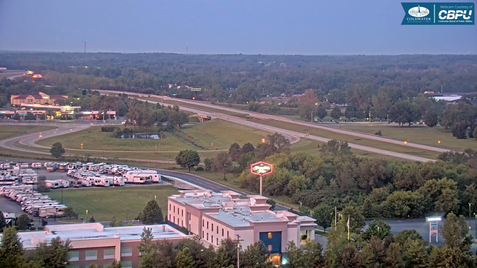 A high-angle, long shot shows a highway, a large parking lot full of RVs, a hotel, and a sprawling area of green trees under a twilight sky.