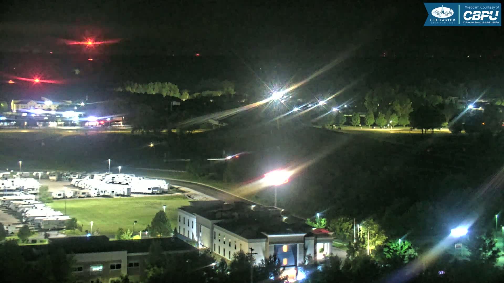 A nighttime aerial view shows a hotel, a large RV park, a roadway with moving vehicles, and a wooded area under clear skies.