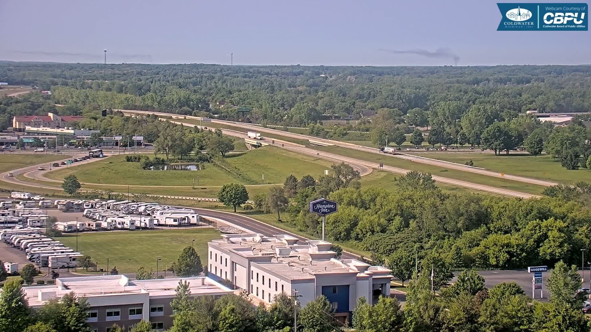 A high-angle, long shot on a clear, sunny day shows a multi-lane highway cutting through a green landscape dotted with buildings, including a hotel and a large RV park.