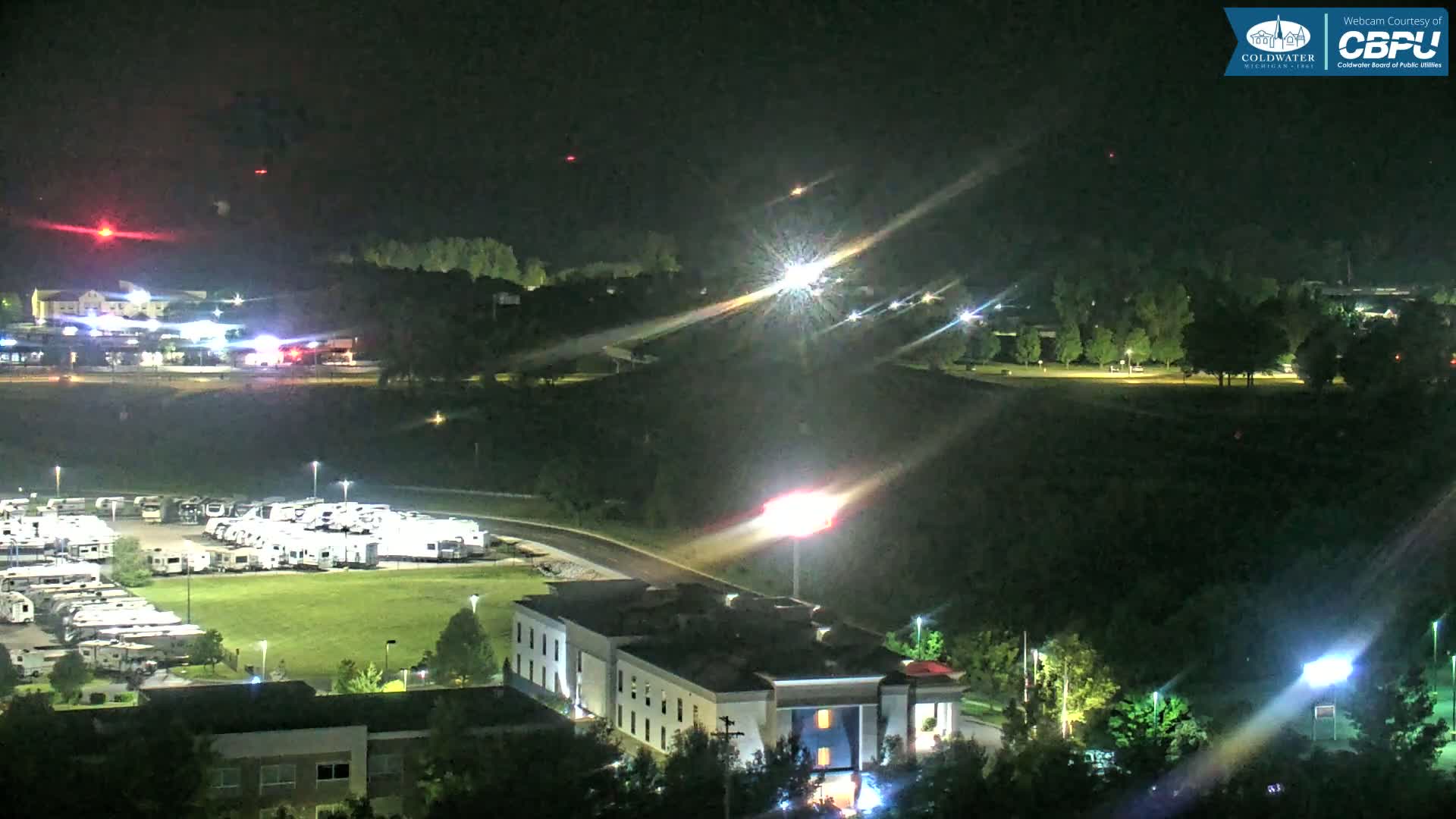 A nighttime aerial view shows a parking lot filled with RVs, a hotel, and trees, with lights illuminating the scene under a clear sky.