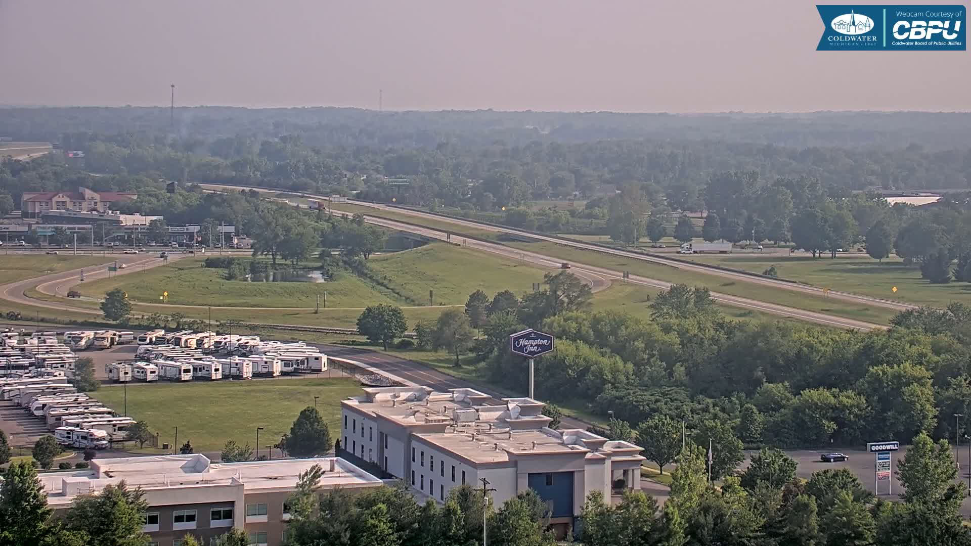 A hazy, summer day shows a highway curving through a landscape of green trees and fields, alongside a Hampton Inn, a large RV park, and other buildings.