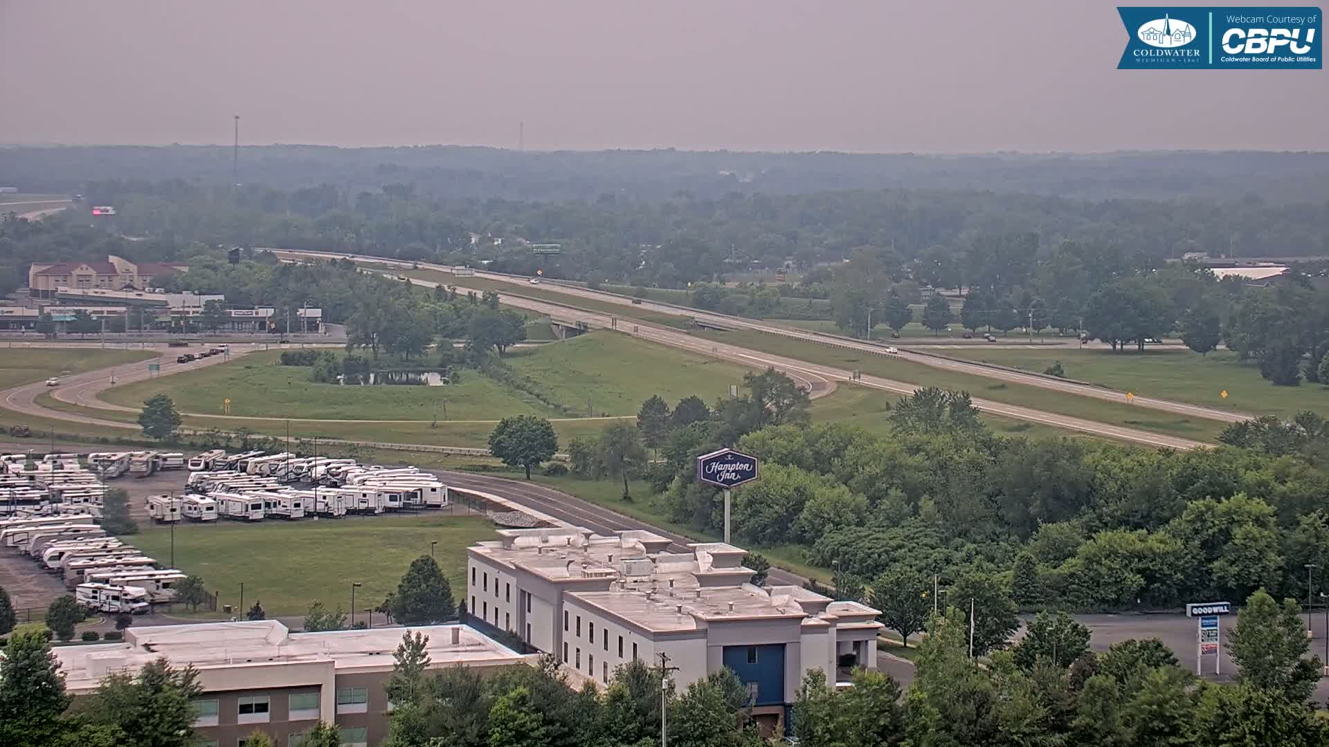 A hazy, overcast day reveals a highway, a large RV parking lot, a Hampton Inn, and surrounding green spaces and buildings.