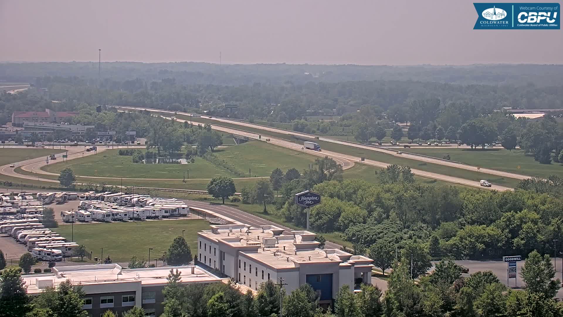 A hazy, summer day reveals a highway, a large RV park, a Hampton Inn, and other buildings and businesses surrounded by green trees and grass.