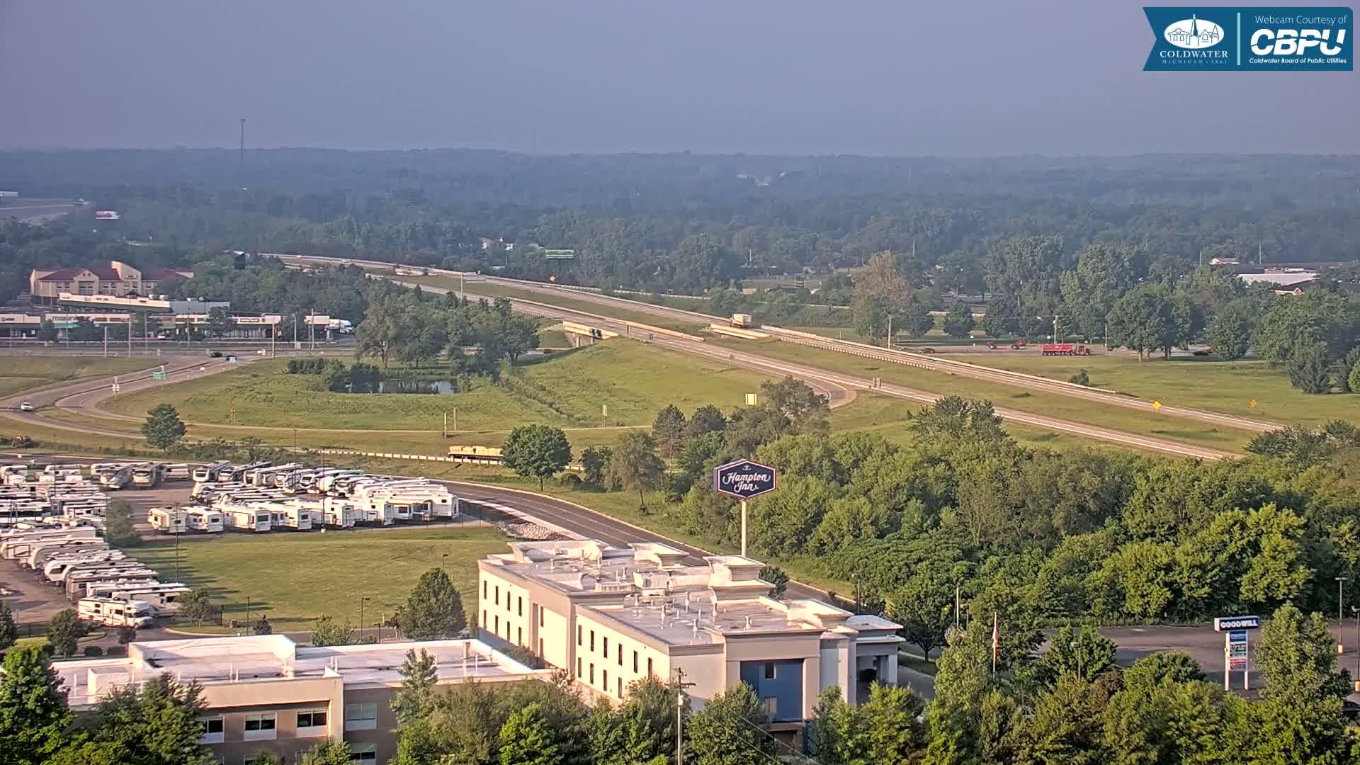 A hazy, sunny day reveals a highway, a large RV park, a Hampton Inn, and surrounding green areas with trees.