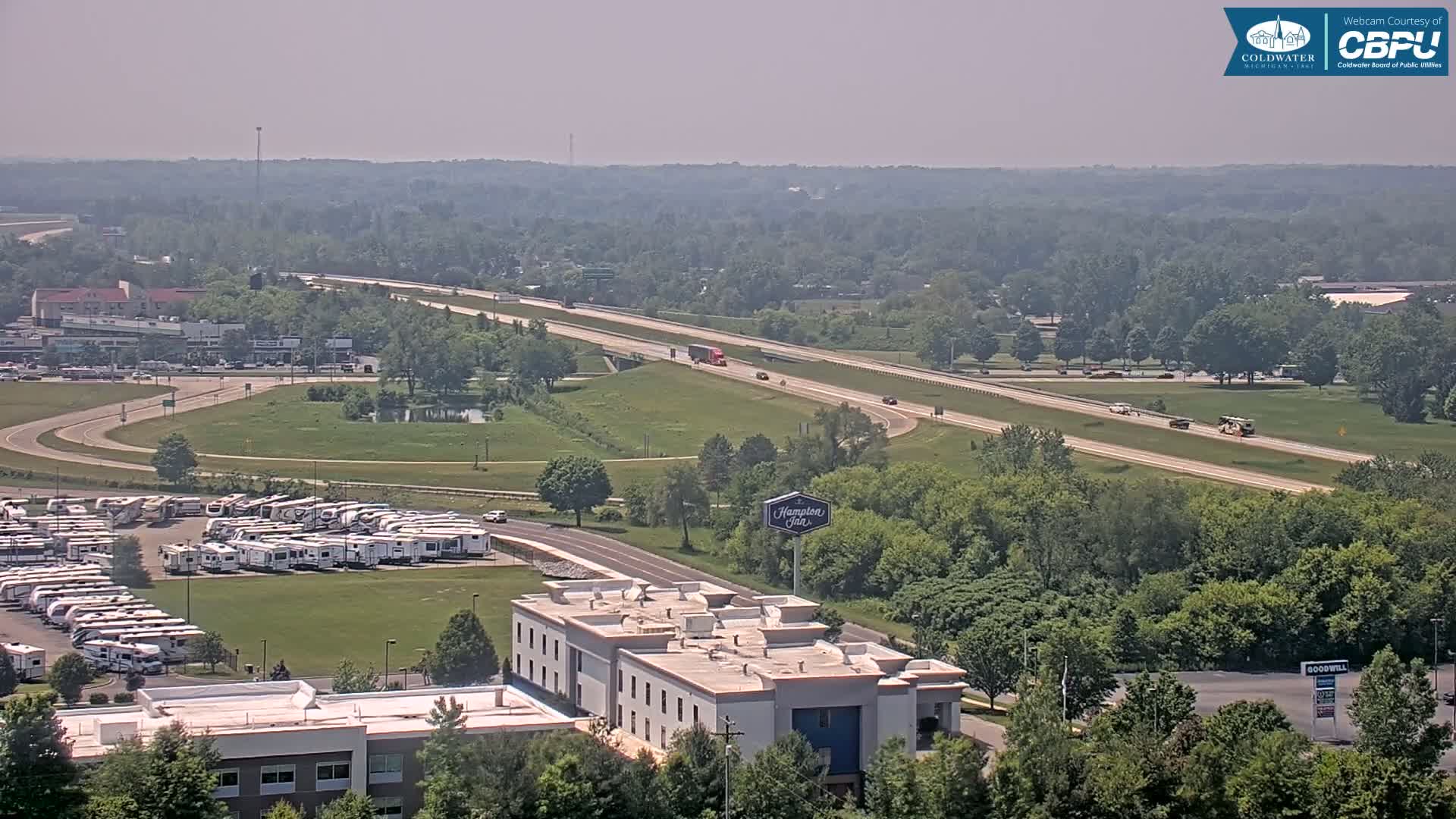 A hazy, sunny day reveals a highway with sparse traffic, a large RV park, and a Hampton Inn amongst green, leafy trees and buildings.