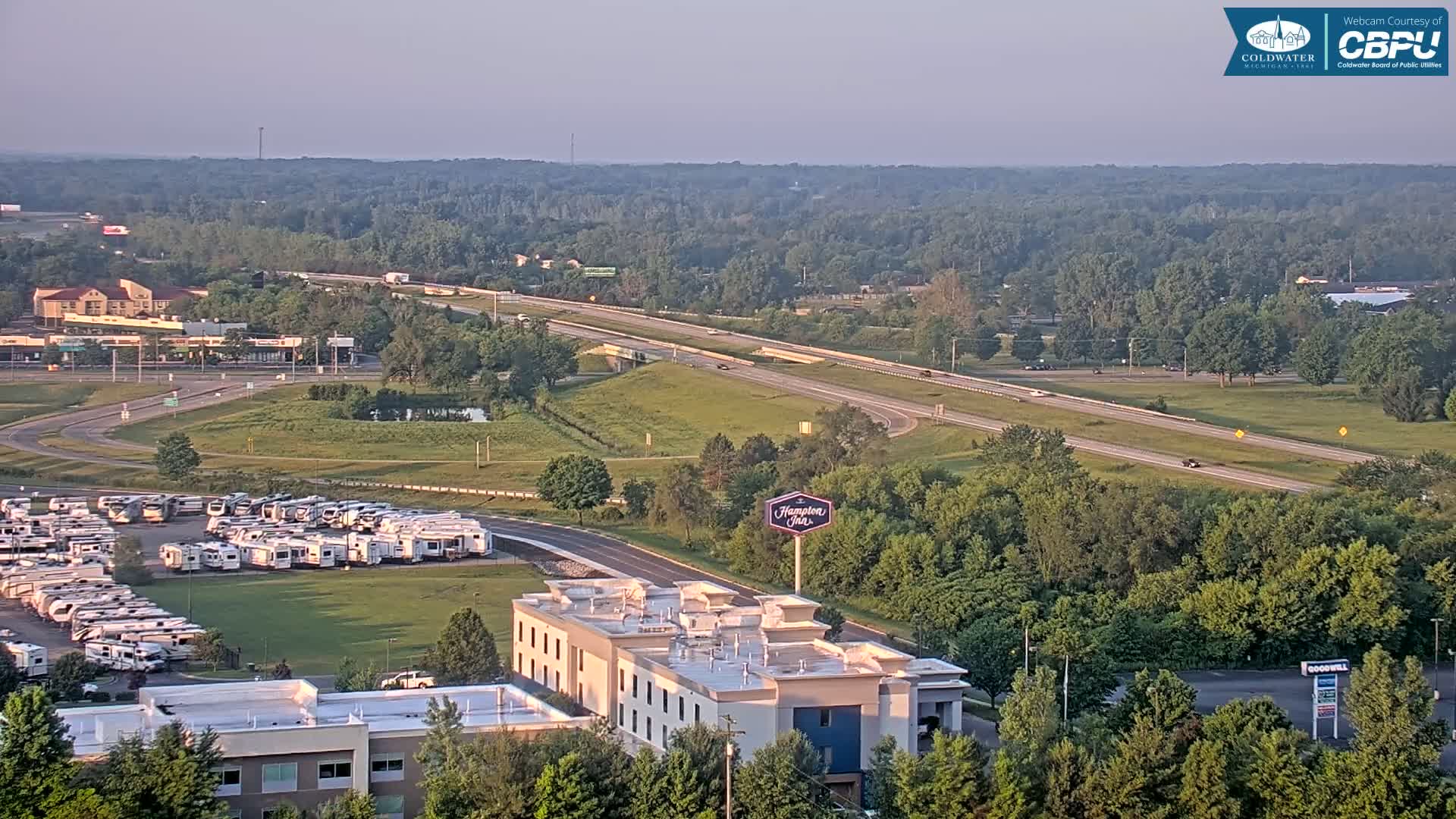 An aerial view shows a highway, a Hampton Inn, a large RV park, and surrounding green spaces under hazy, light conditions.