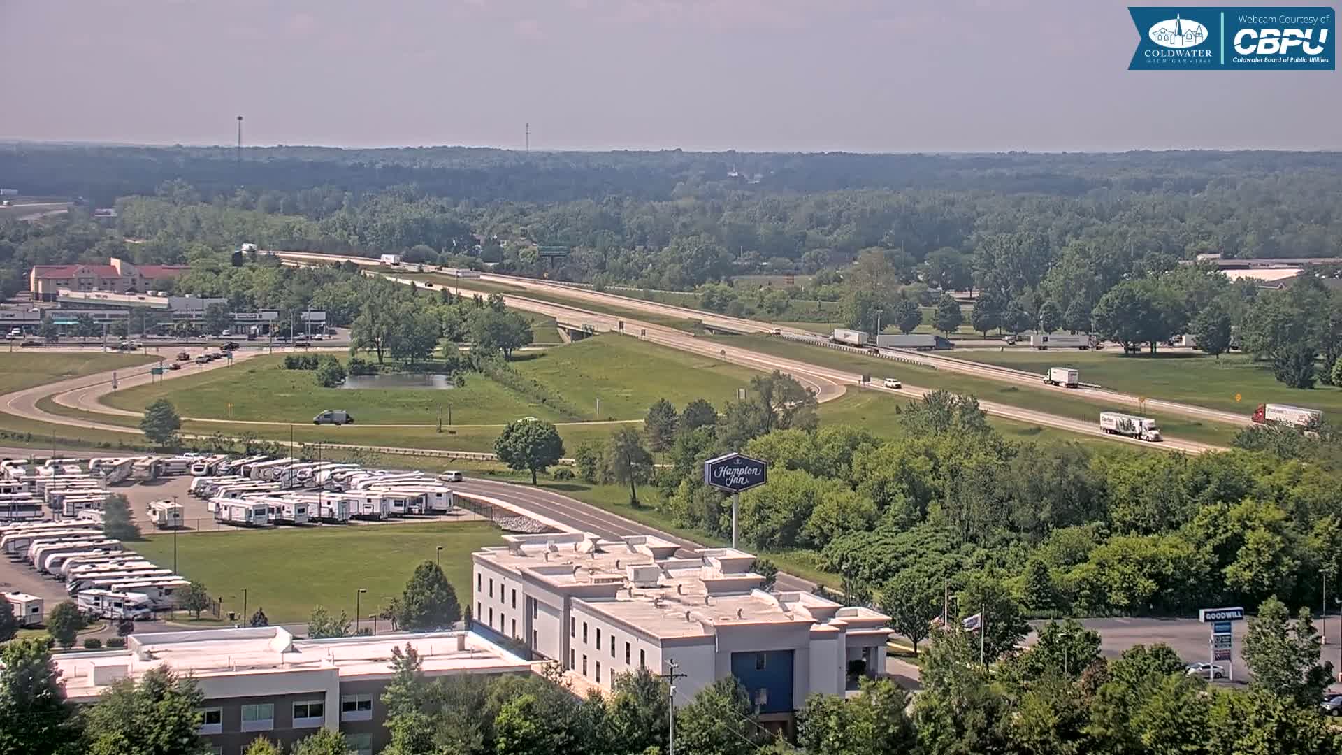 A sunny day reveals a highway with several semi-trucks, alongside a large RV park, a Hampton Inn, and a sprawling wooded area.