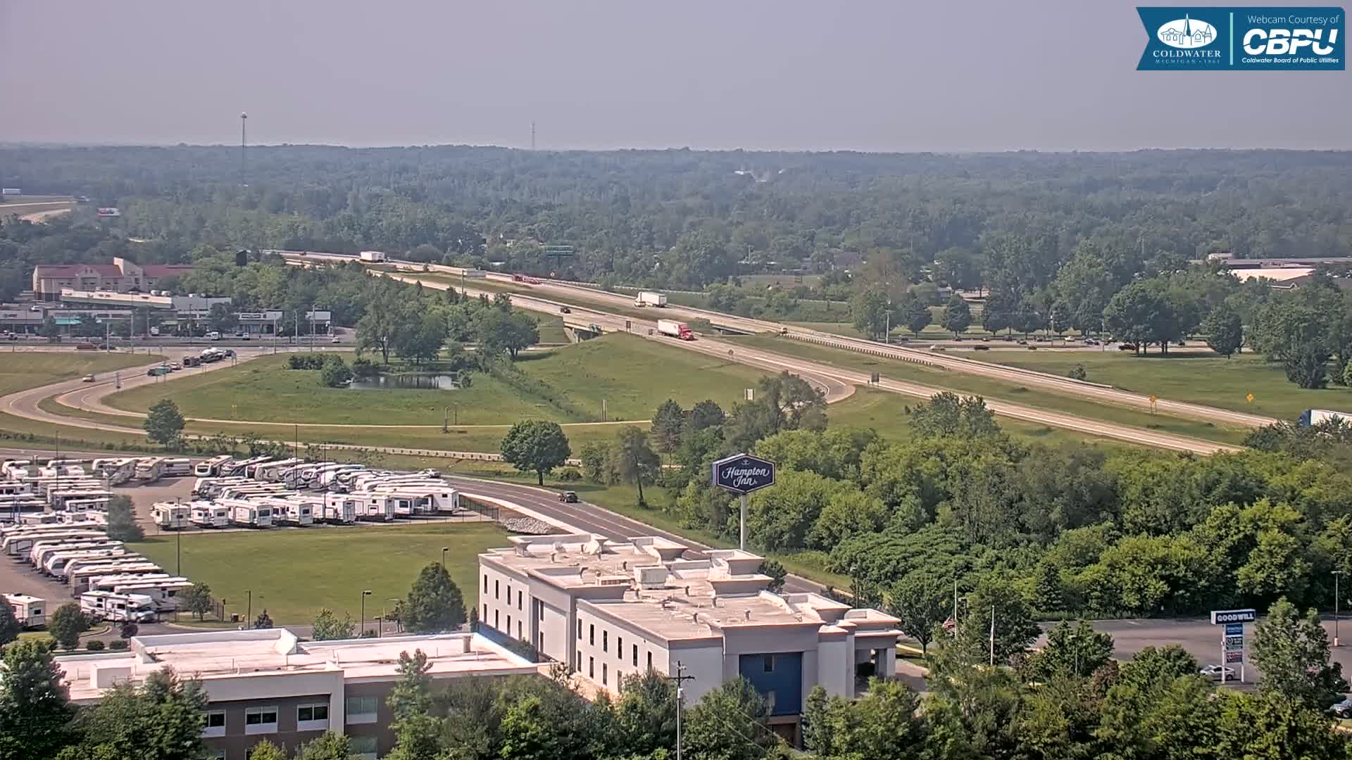 A high-angle, long shot on a hazy, sunny day shows a highway with several semi-trucks, a large RV park, and a Hampton Inn among other buildings and green spaces.