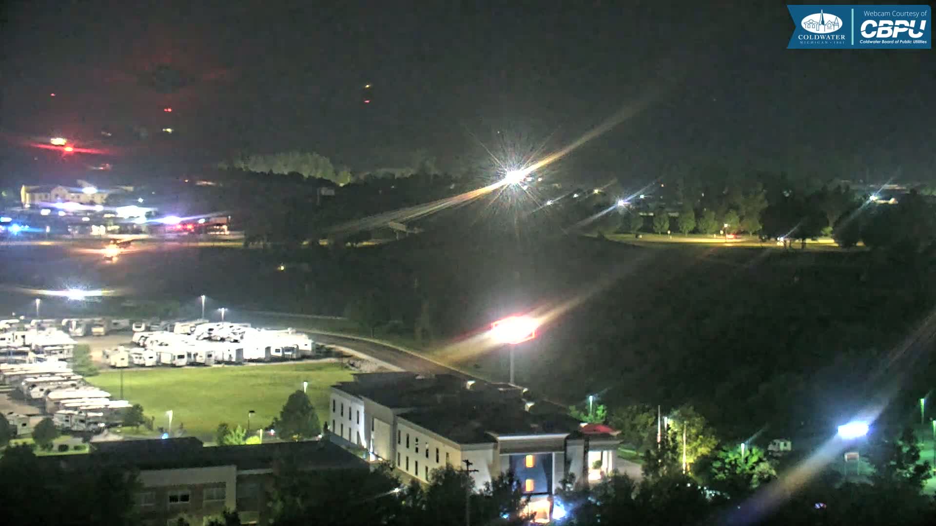A nighttime aerial view shows a parking lot filled with RVs, a hotel, and a roadway, with some lights and distant flashing lights visible under a clear, dark sky.
