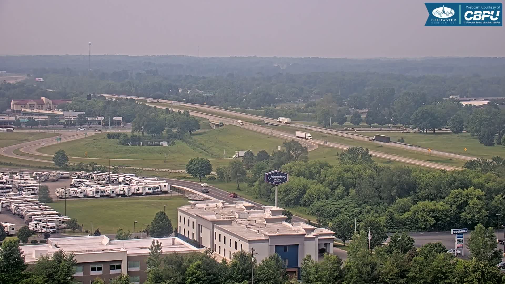 A hazy, summer day reveals a highway with several vehicles, a large RV parking lot, a Hampton Inn, and surrounding green landscape.