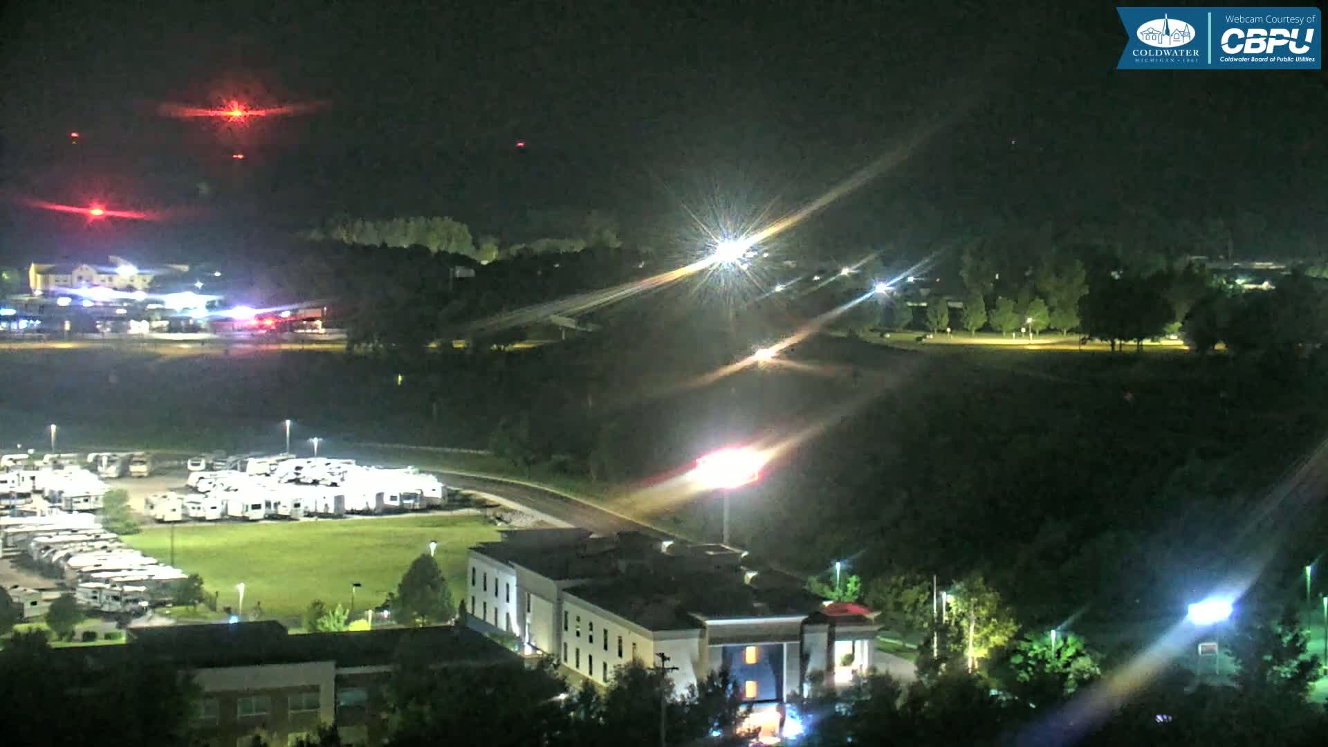A nighttime, high-angle view shows a motel, a large RV park, and a dark, tree-covered hillside under a clear sky with several distant red lights.