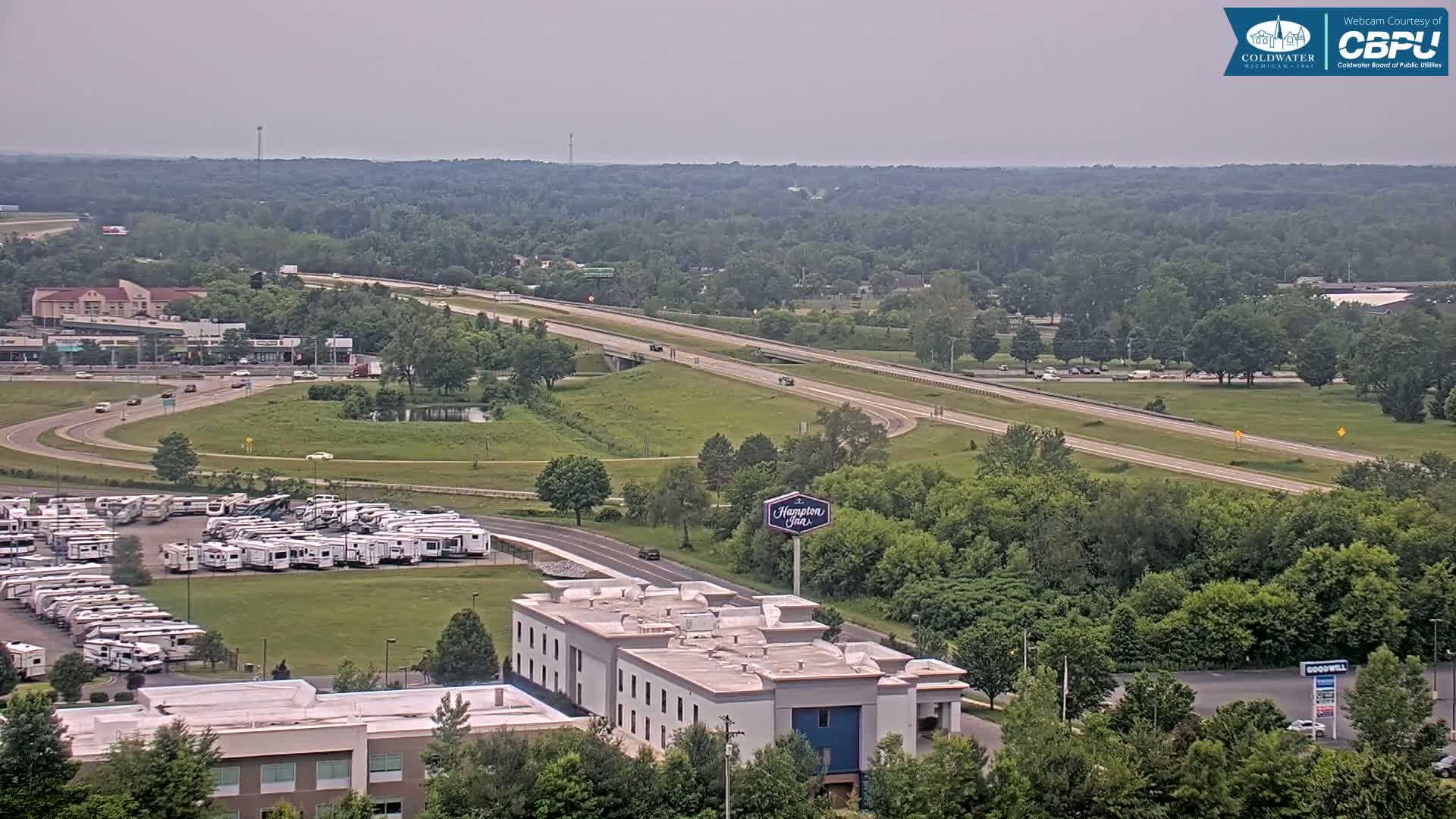 A highway curves through a verdant landscape under a hazy sky, with a large RV park and a hotel visible nearby.