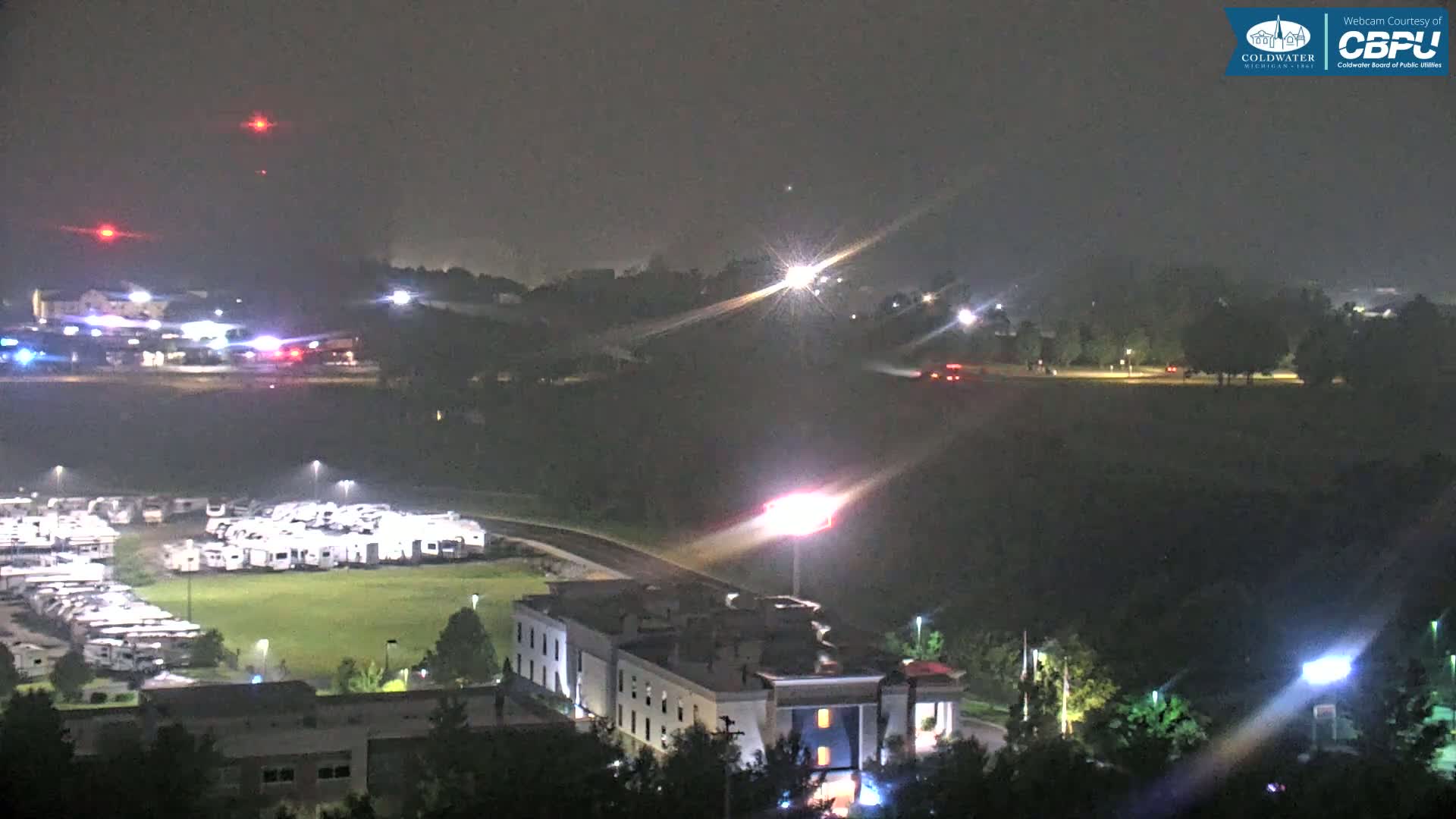 A nighttime view of a landscape featuring a large parking lot filled with RVs, a multi-story building, and some distant lights, possibly from vehicles or emergency responders, under hazy conditions.